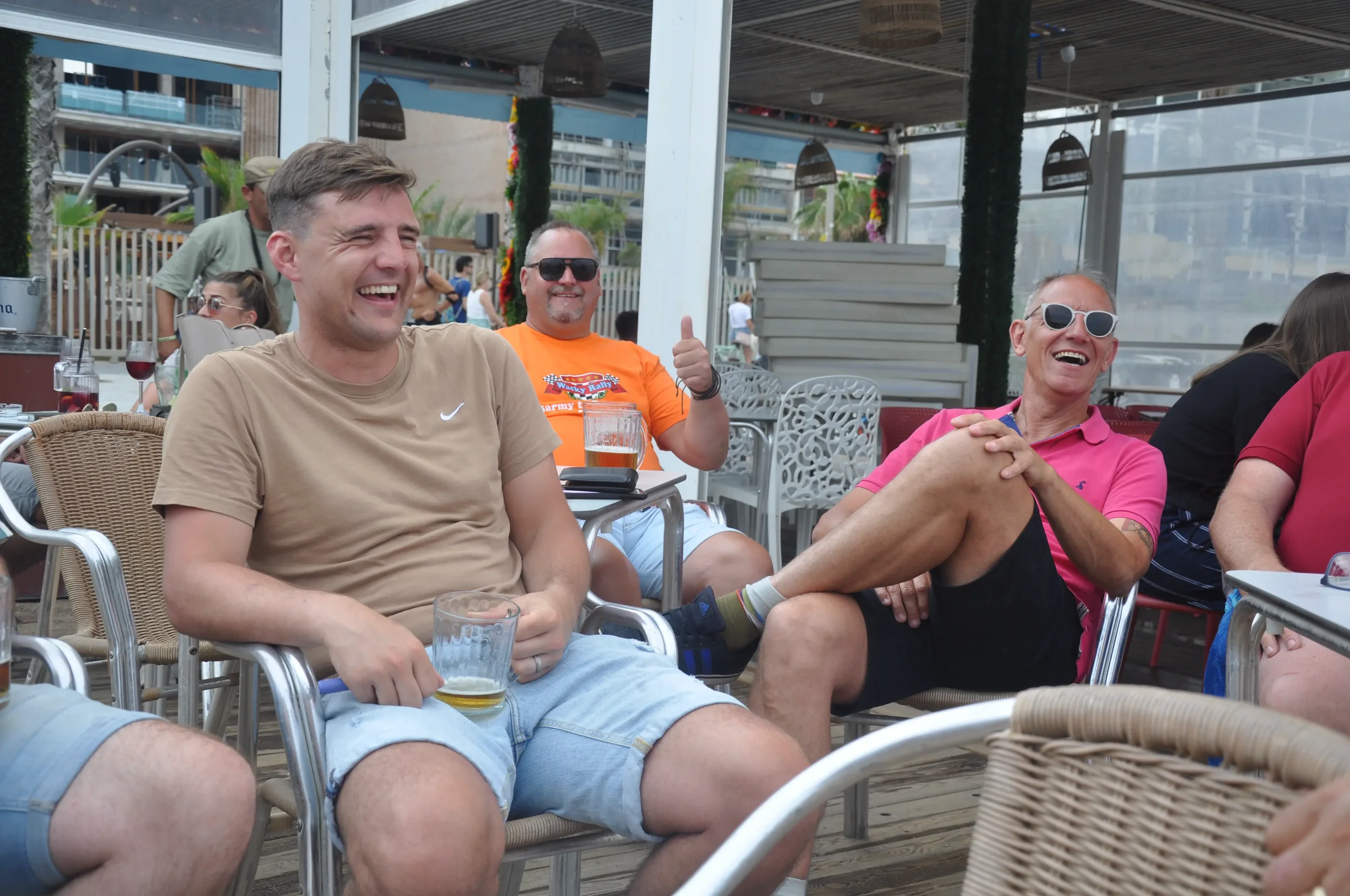 Group of people sitting at an outdoor venue, laughing and smiling. One man in a tan shirt has a beer, and another man in a pink shirt and sunglasses is laughing with crossed legs. Others are visible in the background.