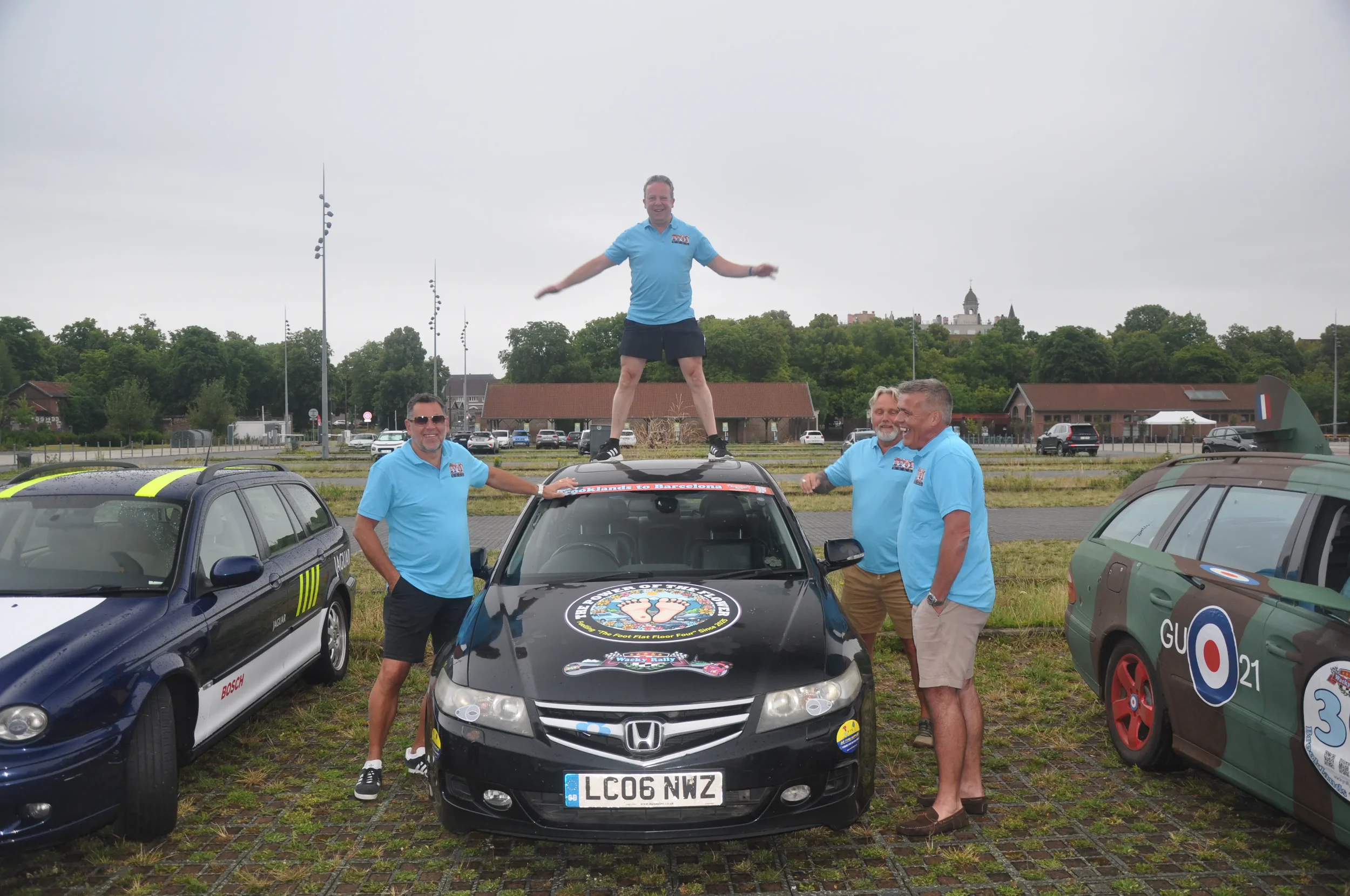 Five men in blue shirts standing around and on a black Honda Civic parked between two cars, with one man standing on top of the car on a jumping pose. The cars have various decals and stickers, and the scene appears to be in a parking lot on a cloudy