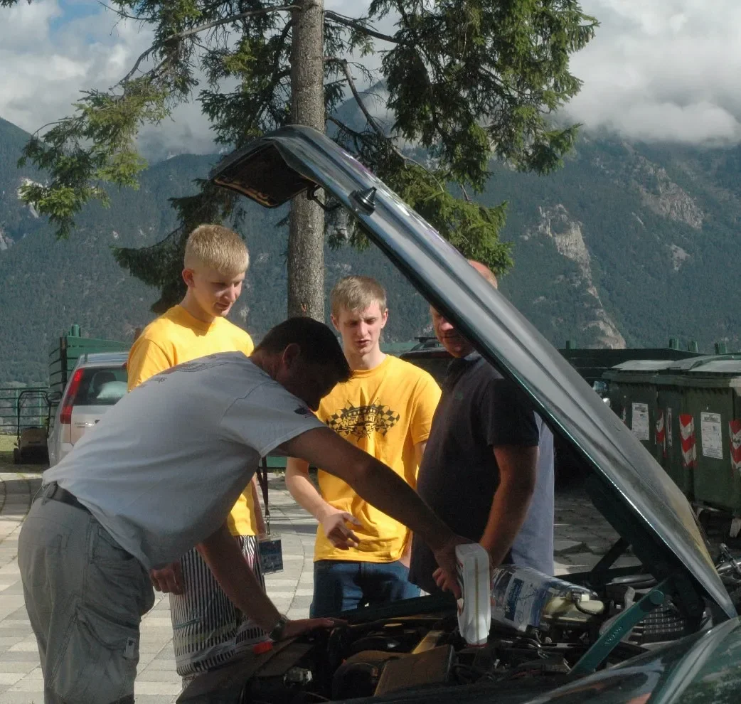 Four men are looking under the open hood of a car. Three of them are young men wearing yellow T-shirts, and the other man is older, wearing a dark shirt. They are outdoors in a mountainous area with trees and clouds in the background.