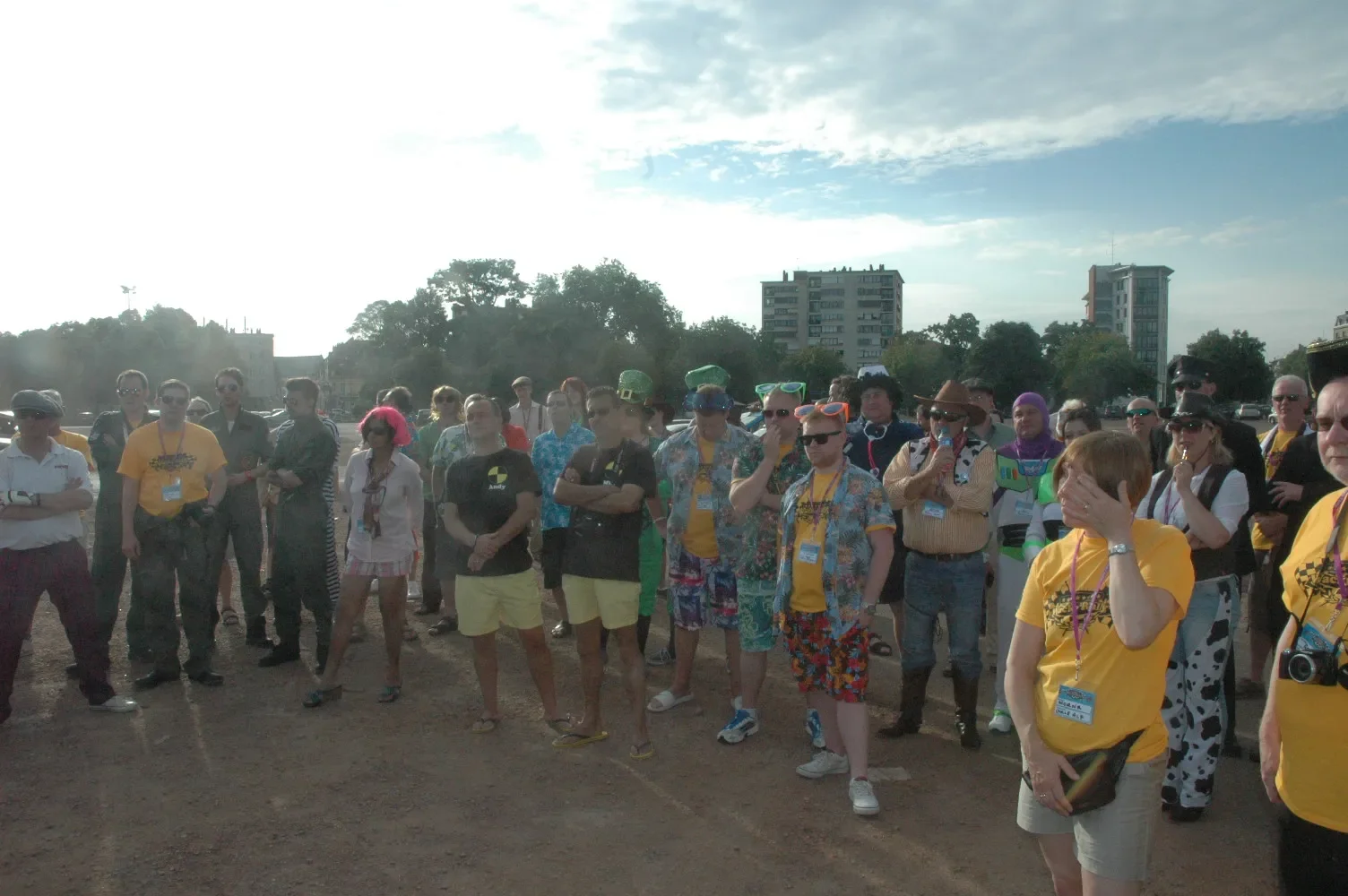 Group of people gathered outdoors on a sunny day, some wearing colorful costumes and accessories, with a cityscape of buildings and trees in the background.