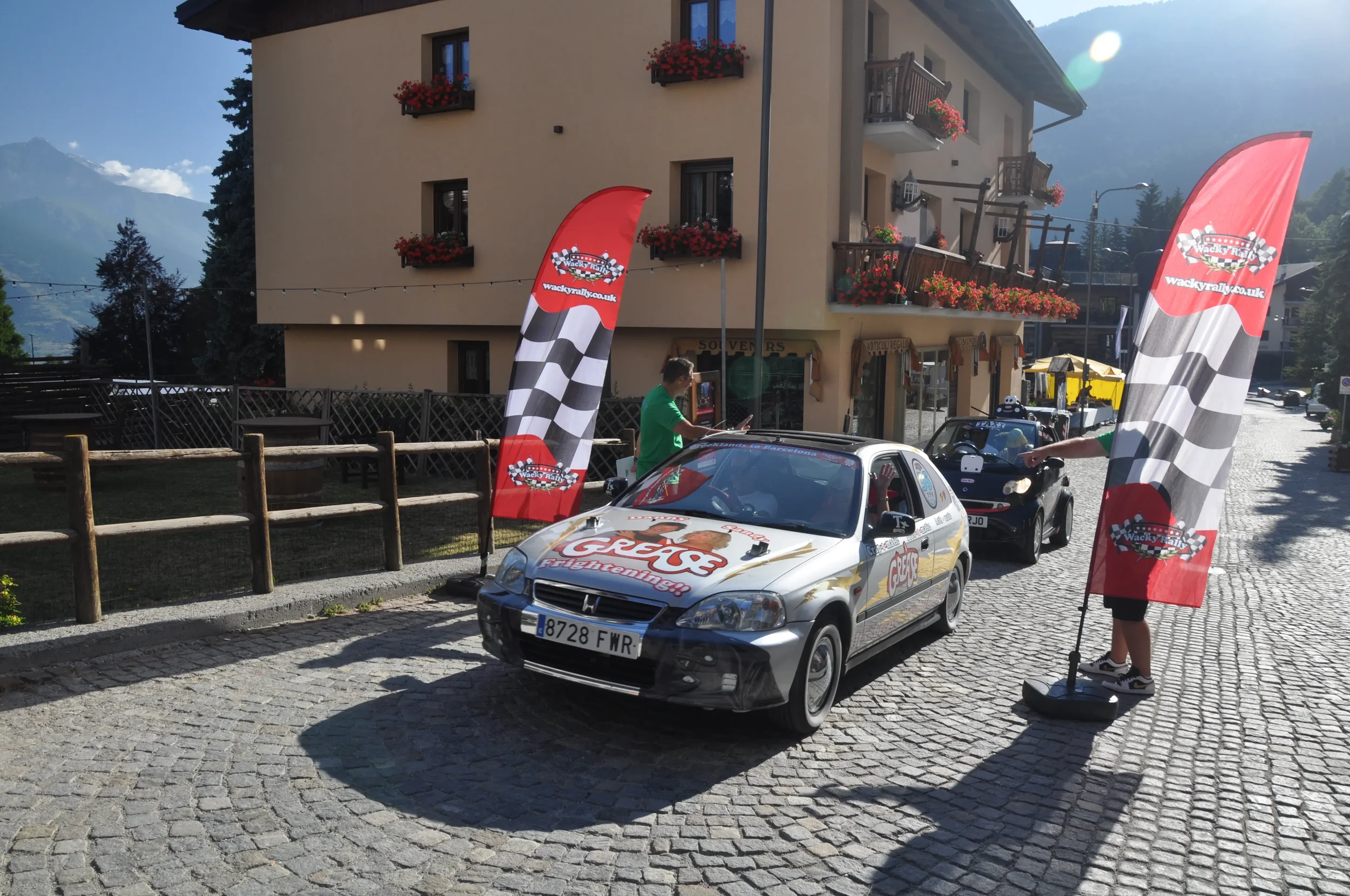 A rally car is parked on a cobblestone street in front of a building with flower boxes on the balconies. Two flags with checkered patterns and the URL wacky rally.co.uk are displayed next to the car. Two people are near the car, one leaning in throug