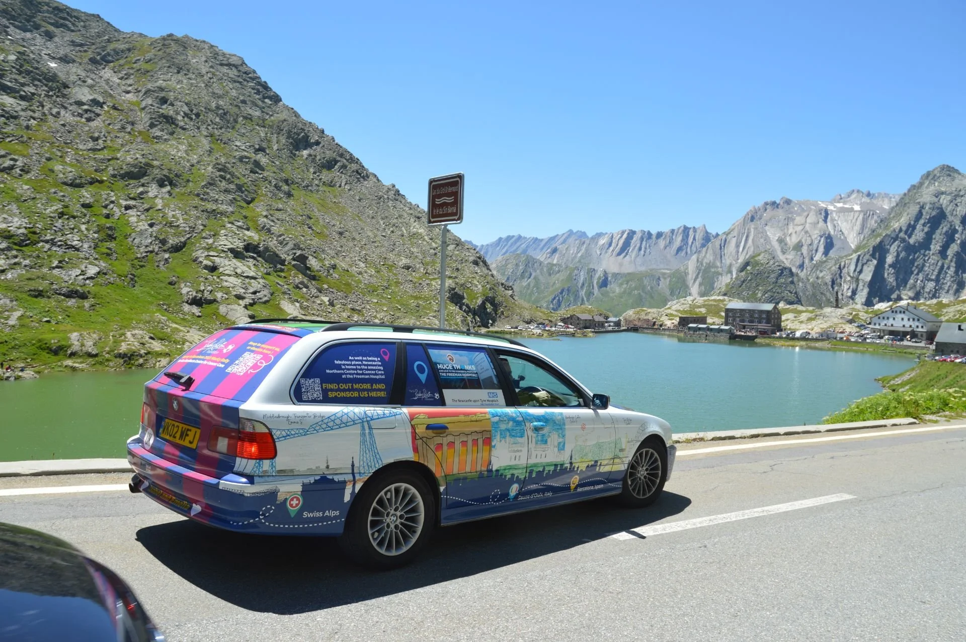 A colorful car driving highway near a lake surrounded by mountains and a small town in the distance.