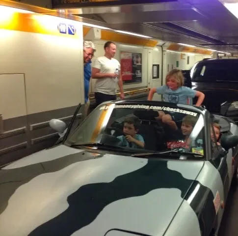 Children and adults at an airport baggage claim, with some children sitting in a small black and white toy car.