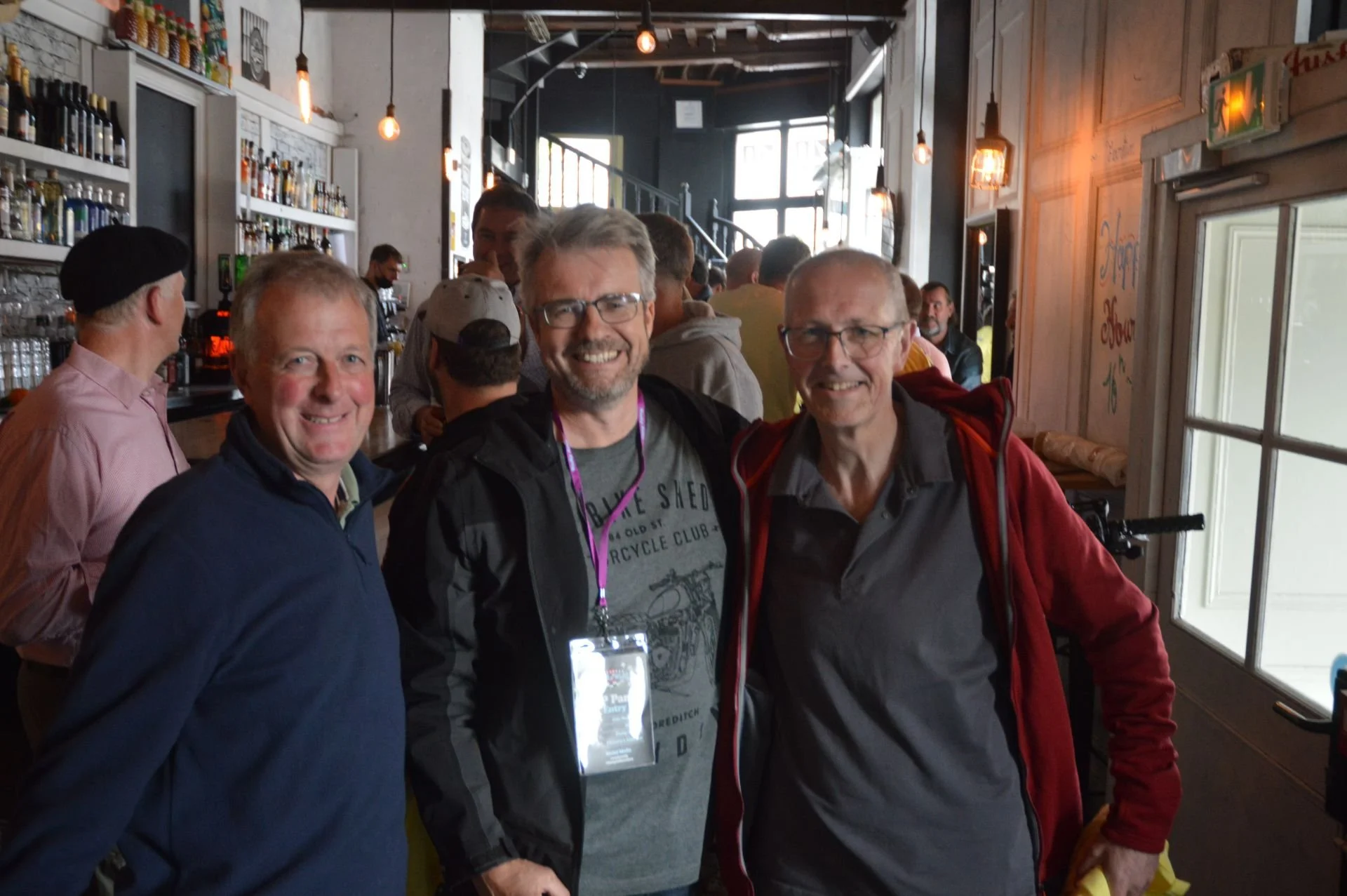 Three smiling men standing inside a busy bar or cafe, with bottles on shelves and other patrons in the background.