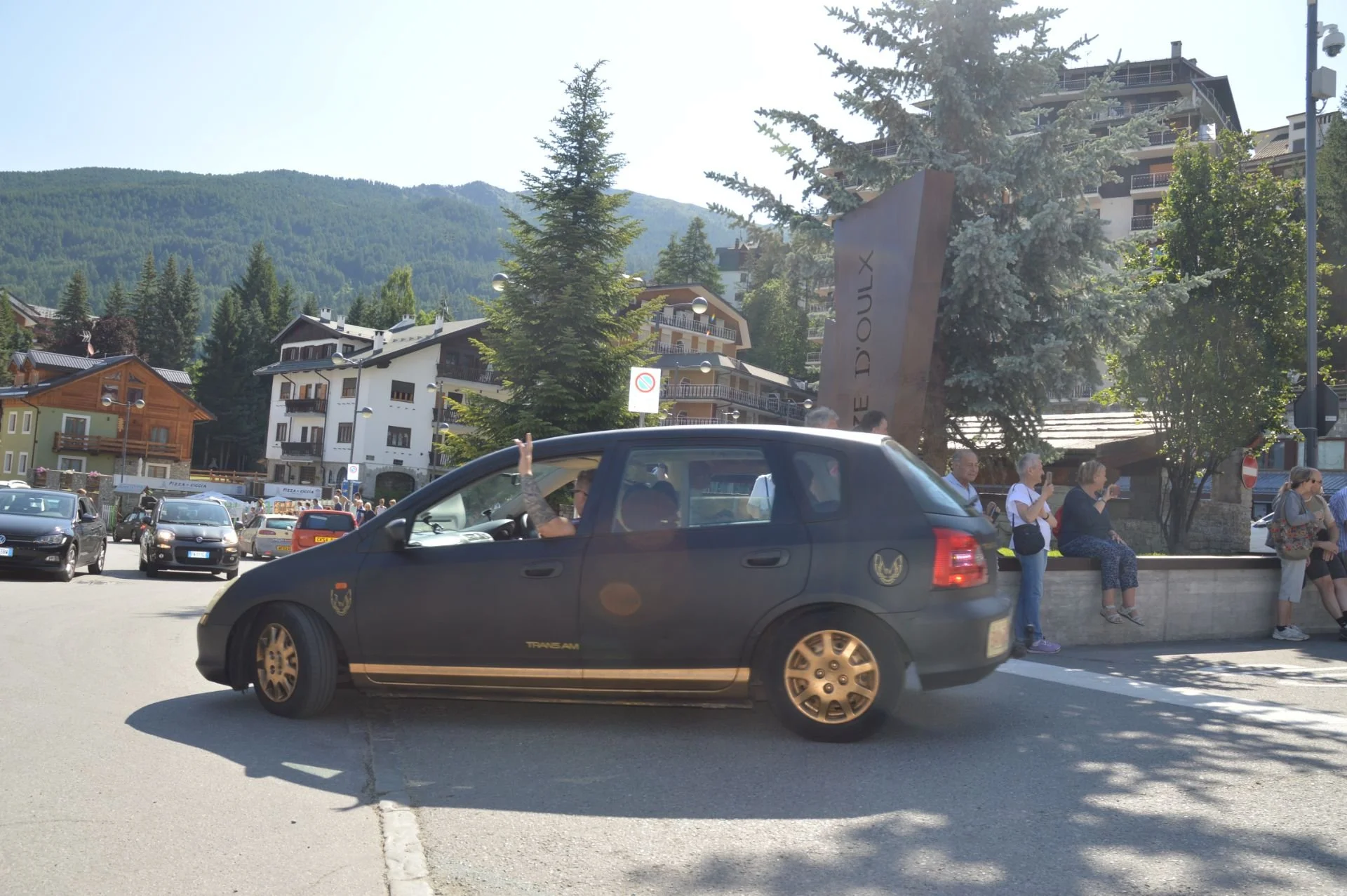 A person in a dark blue police or military vehicle waving from the front passenger window, with a group of people sitting on a stone ledge and standing nearby, in a mountain town with trees and buildings in the background.