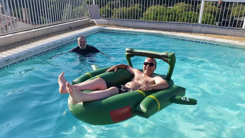 Two men enjoying a swimming pool; one is sitting on an inflatable raft and the other is standing in the pool.
