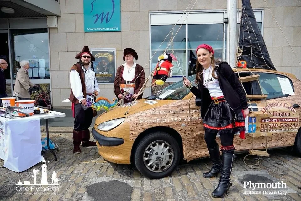 Three people in pirate costumes standing next to a car decorated like a pirate ship at an event outside Mayflower Museum, with a table of items and a woman holding a pirate sword.