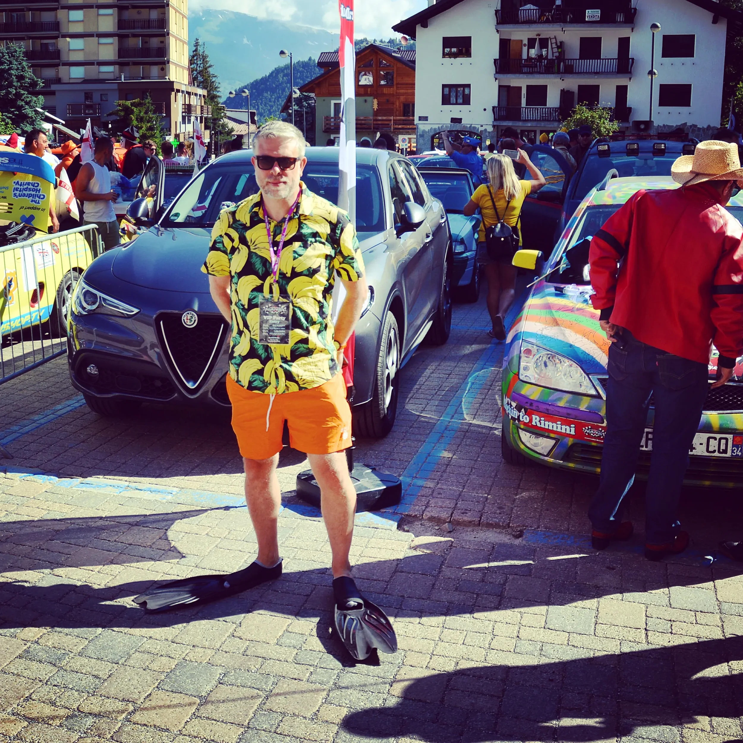A man in a yellow banana print shirt and orange shorts stands on a paved street wearing diving fins, with a crowd, cars, and buildings in the background at an outdoor event.