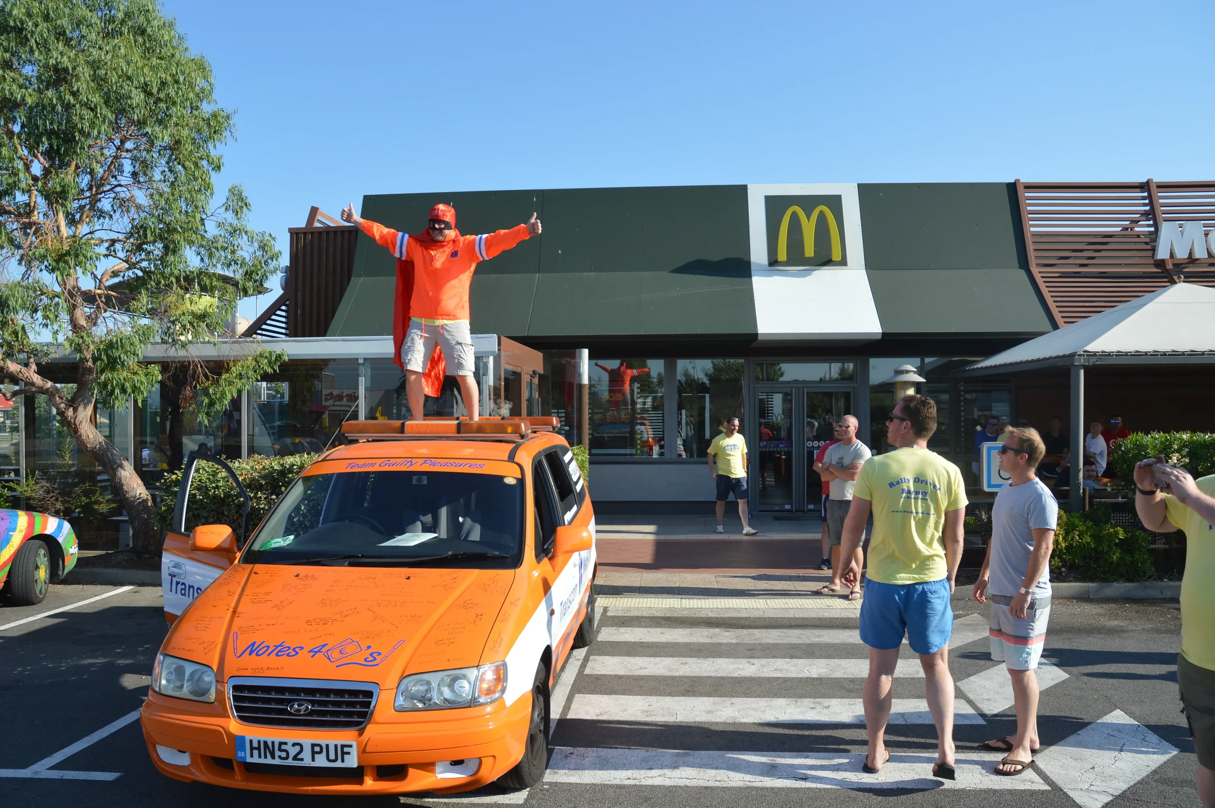 A group of people gathered outside a McDonald's restaurant on a sunny day, with a man standing on an orange car giving a thumbs-up, dressed in a red superhero cape and baseball cap.