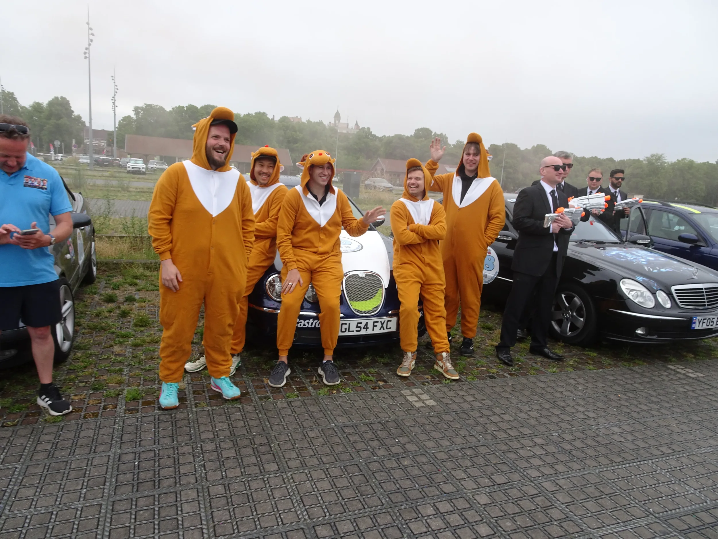 Group of people, some dressed in lion costumes and others in black suits with toy guns, standing in a parking lot with cars and a cloudy sky in the background.