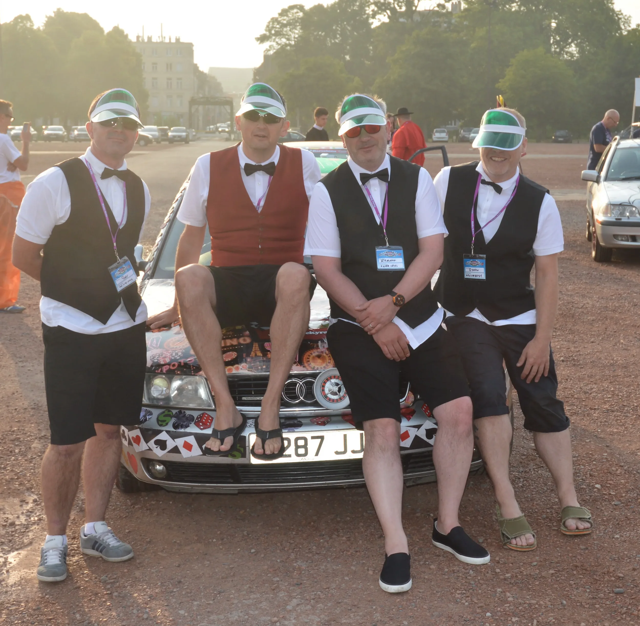 Four men dressed in tuxedo vests, shorts, and summer footwear, standing and sitting around a decorated car, wearing visors and sunglasses, at an outdoor gathering during sunset.