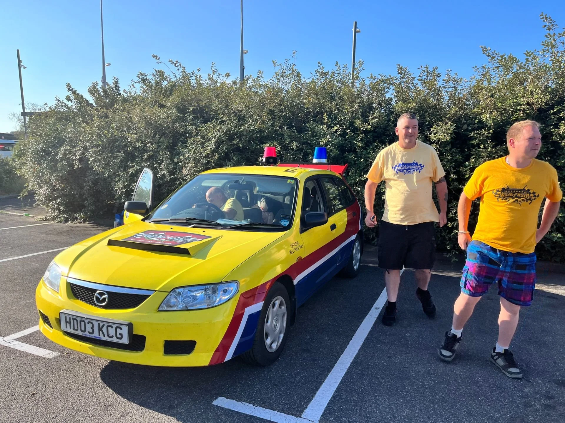 Yellow police-themed car with red and blue stripes and emergency lights, parked in a parking lot with two men walking nearby, one wearing a yellow shirt and the other wearing a beige shirt, with bushes and tall light poles in the background.
