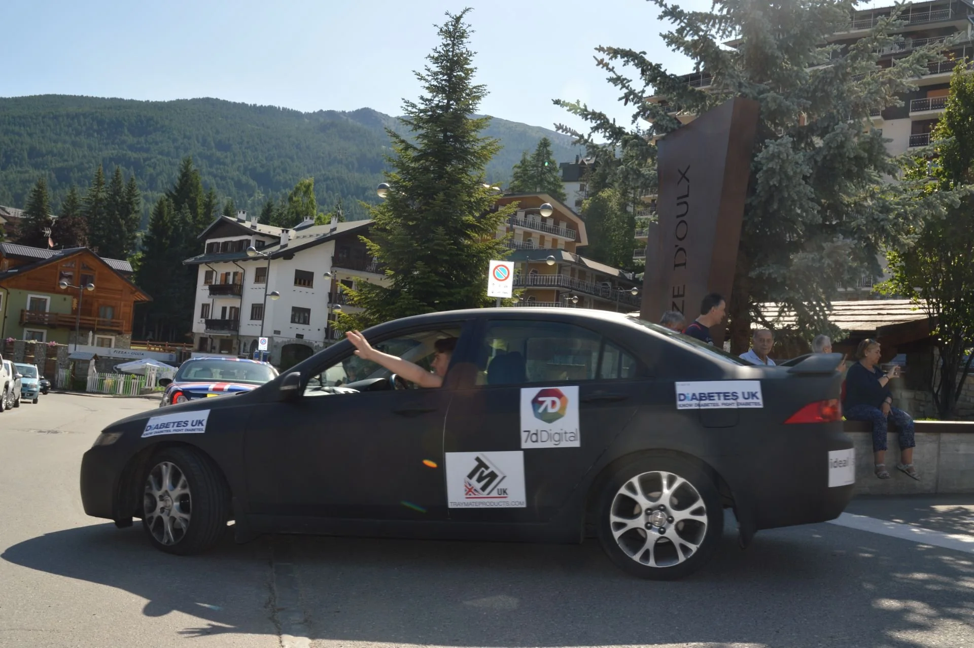 A black car with multiple stickers parked in a mountainous town. People are sitting and standing nearby, with multi-story buildings and trees in the background.