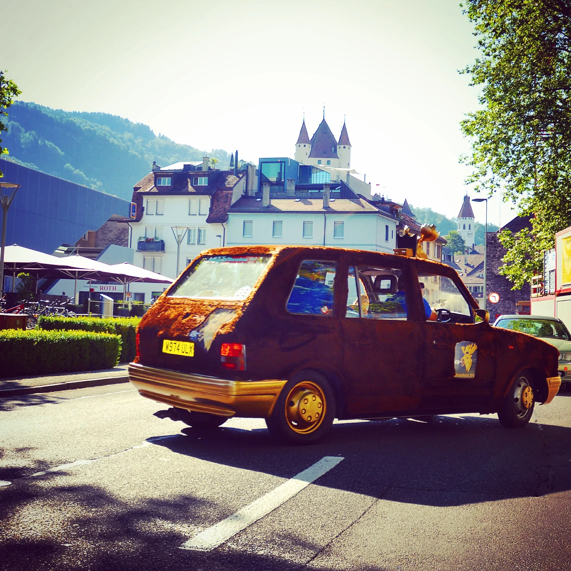 Rusty toy car with a ginger cat atop it, parked on a street with buildings and a castle hill in the background.