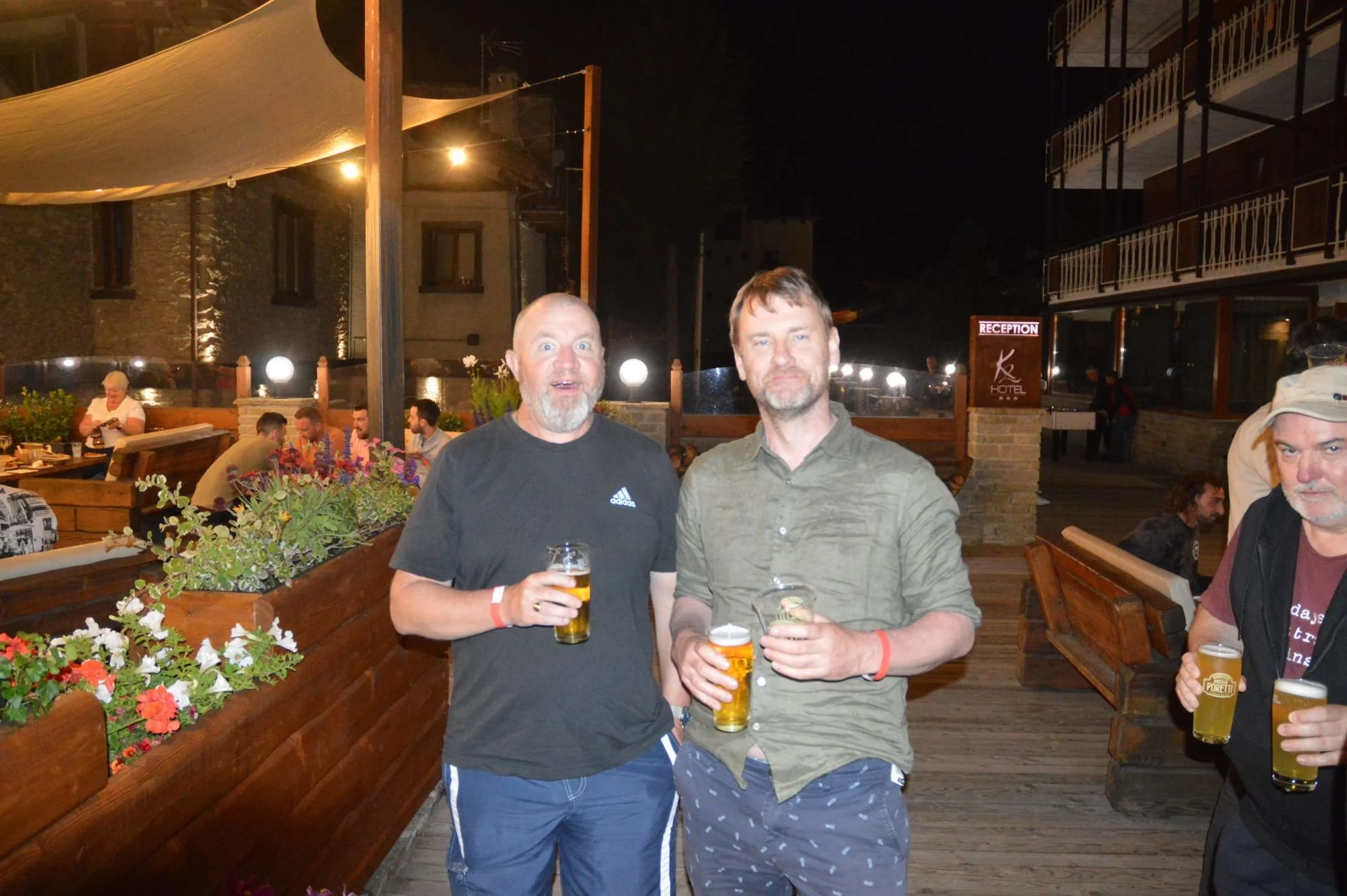 Two men standing outdoors at night, holding beers, with a restaurant patio and other people sitting at tables in the background.