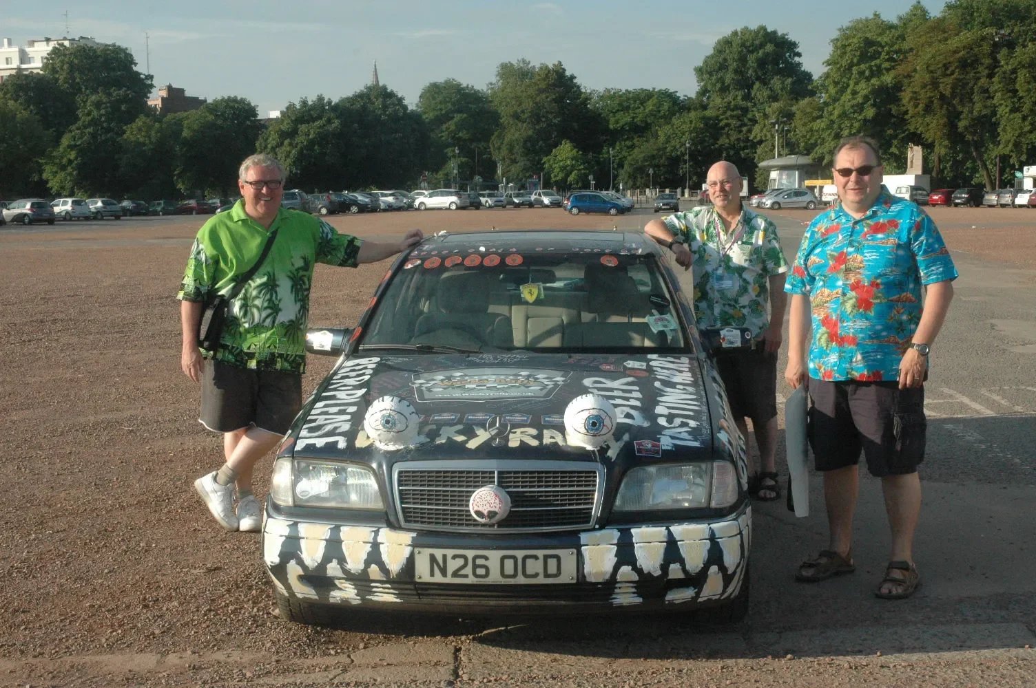 Three men in colorful Hawaiian shirts standing next to a decorated Mercedes-Benz car in a parking lot with trees and other cars in the background.
