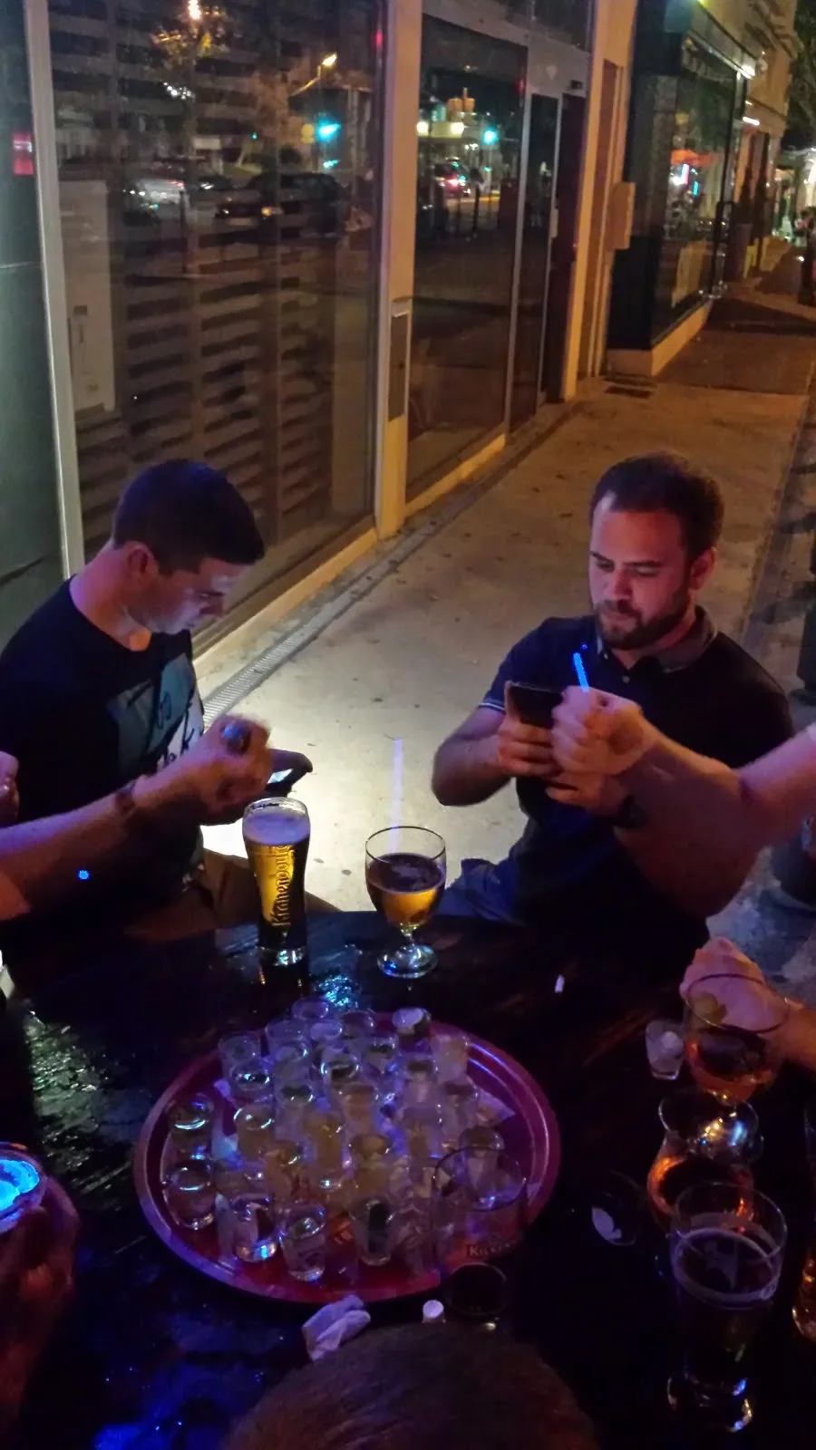 Two men sitting at a table outside a bar or restaurant at night, drinking beer, with a tray of shot glasses and others drinks on the table.