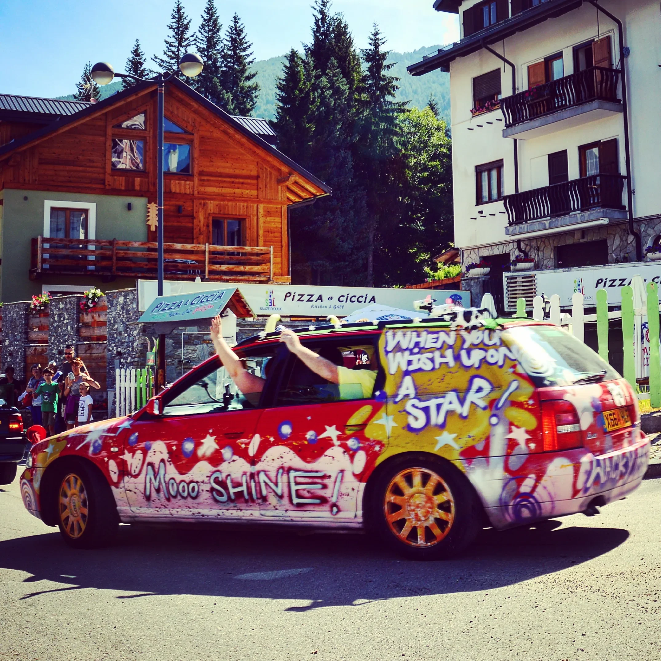 A red car decorated with colorful graffiti, including phrases like 'Mooo…SHINE!' and 'When you wish upon a star!', driving down a street with people watching in the background, and scenic houses with trees behind.
