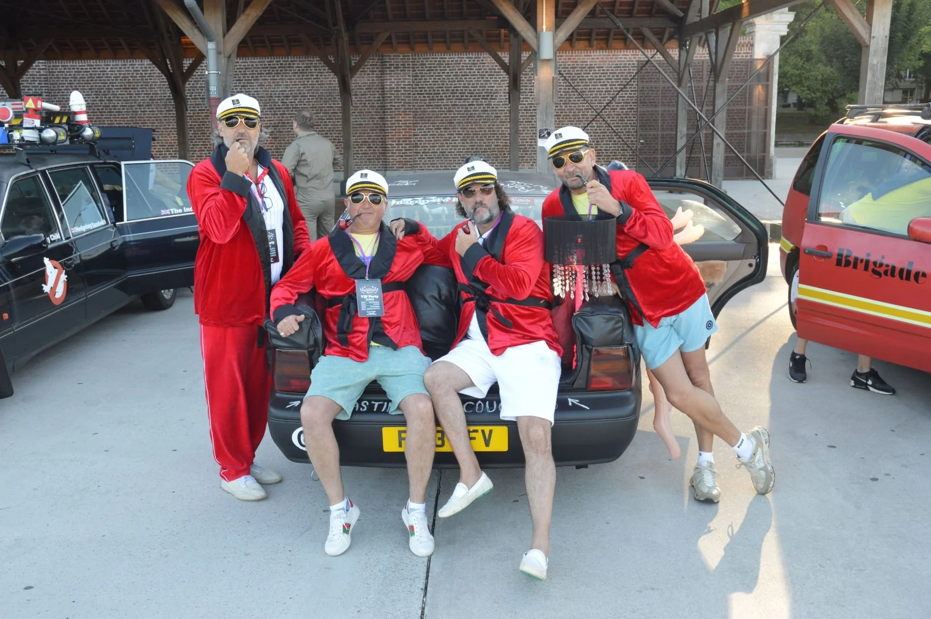 Four men dressed as Ghostbusters wearing red jackets and white hats, sitting and standing on the back of a car, with one man holding a mini proton pack, in front of a brick and wooden structure, with other vintage vehicles and people in the backgroun