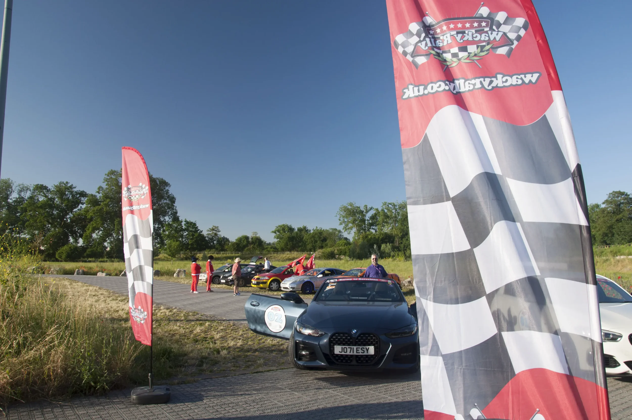 Outdoor car gathering with racing cars and flags, people walking around under clear blue sky.