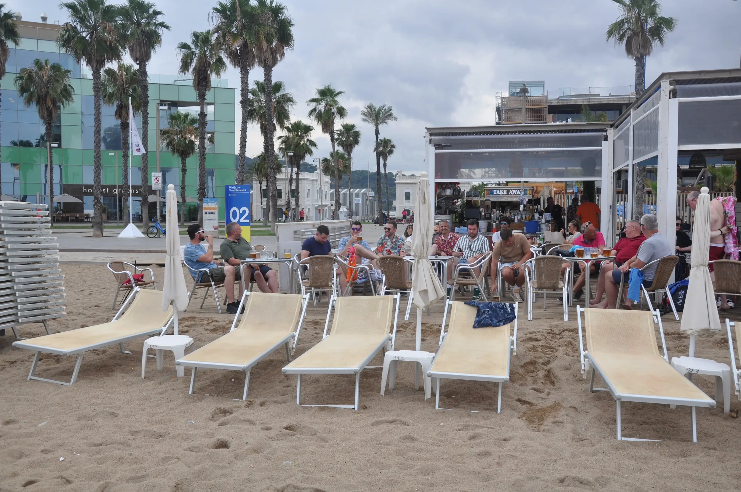 People sitting at tables and lounging on beach chairs at an outdoor beachside cafe, with palm trees, modern buildings, and a cloudy sky in the background.