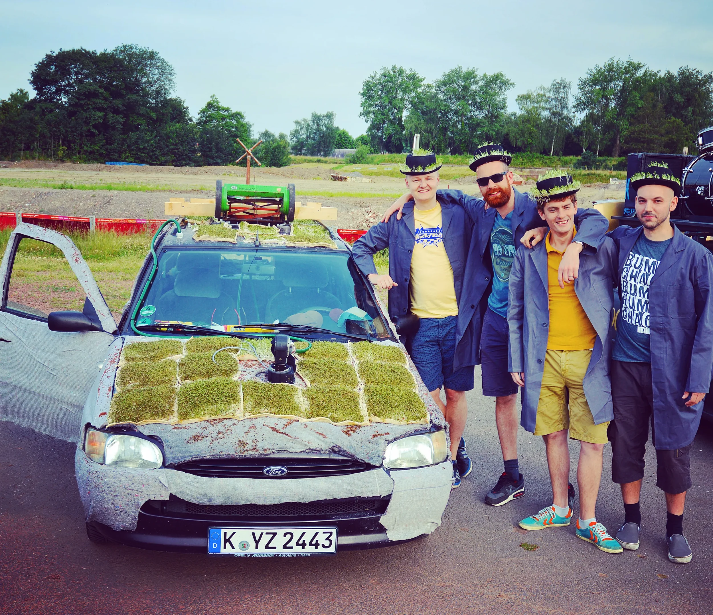 A group of five people posing outdoors next to a car covered in moss and grass, with elaborate decorations, wearing matching hats and casual clothing.