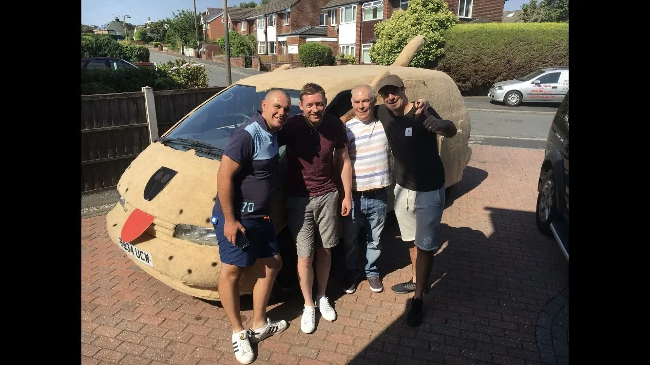 Four men standing beside a car covered with a tan-looking material, making it resemble a giant piece of bread or a pastry. They are outdoors on a driveway, smiling and posing for the photo.