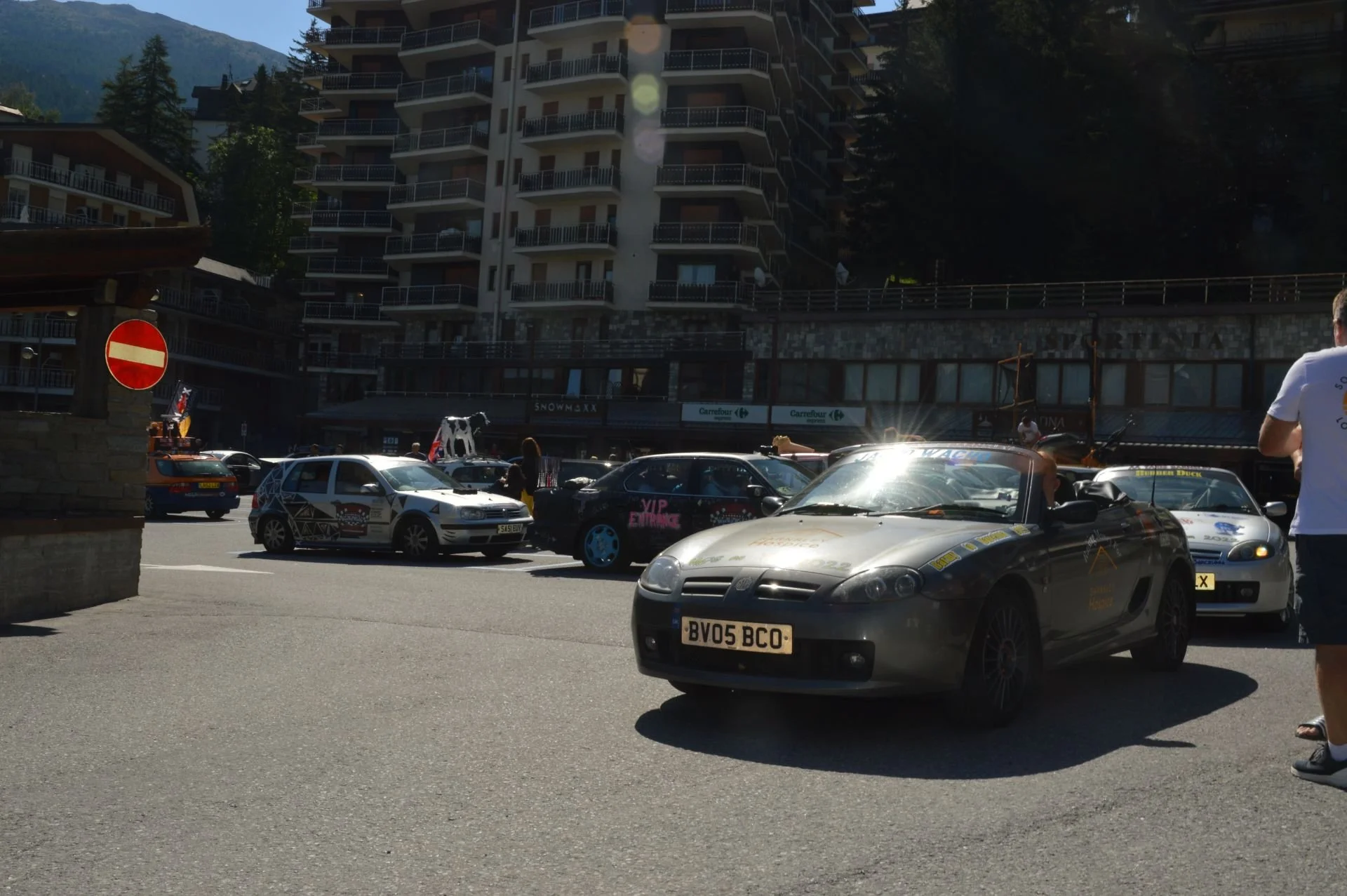 Multiple cars parked in front of a tall building with balconies, with people walking around, and a mountain in the background. The scene appears to be at a car event or gathering.