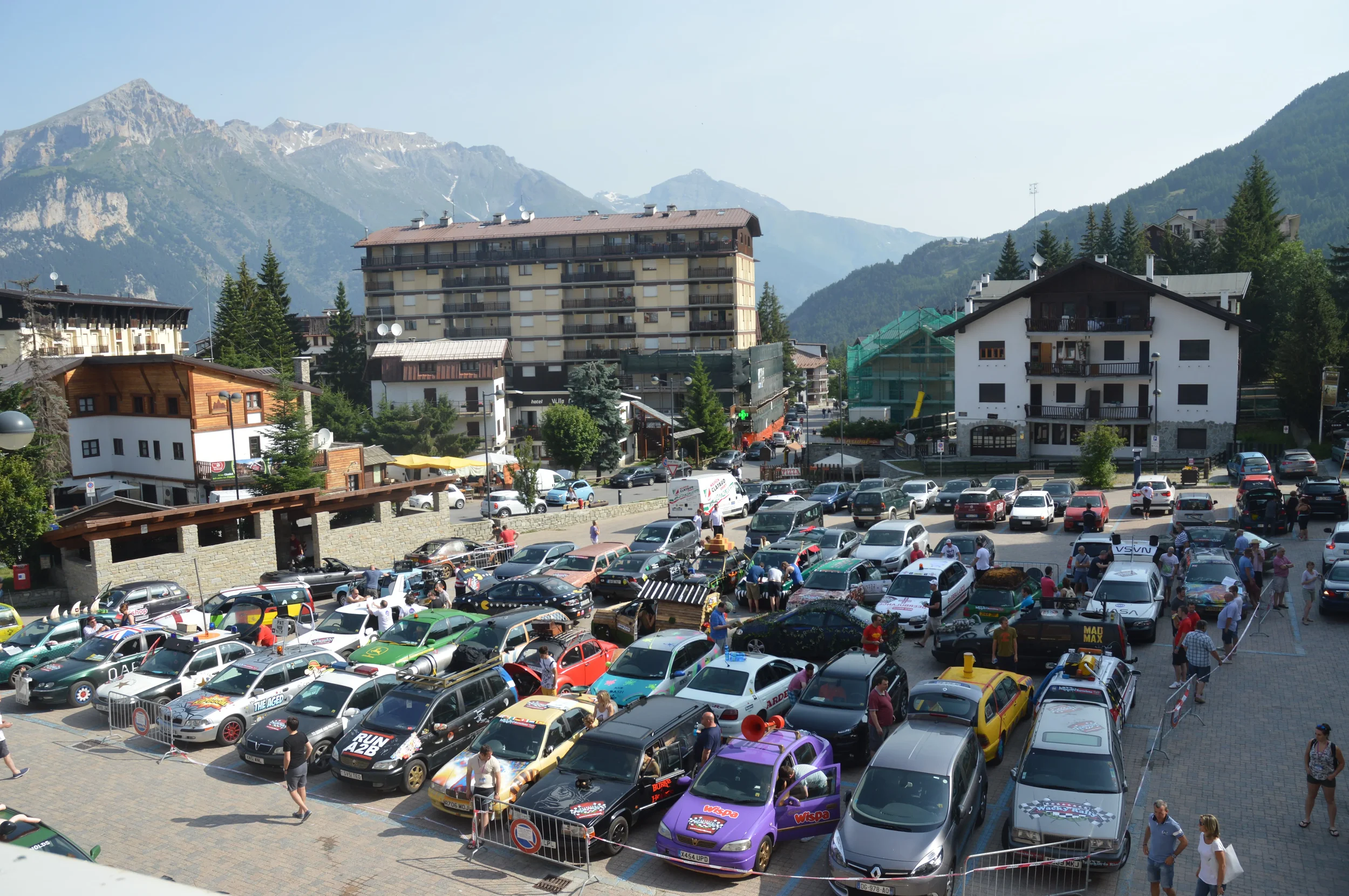 A busy mountain town parking lot filled with cars and people, surrounded by buildings and green trees, with mountains in the background.