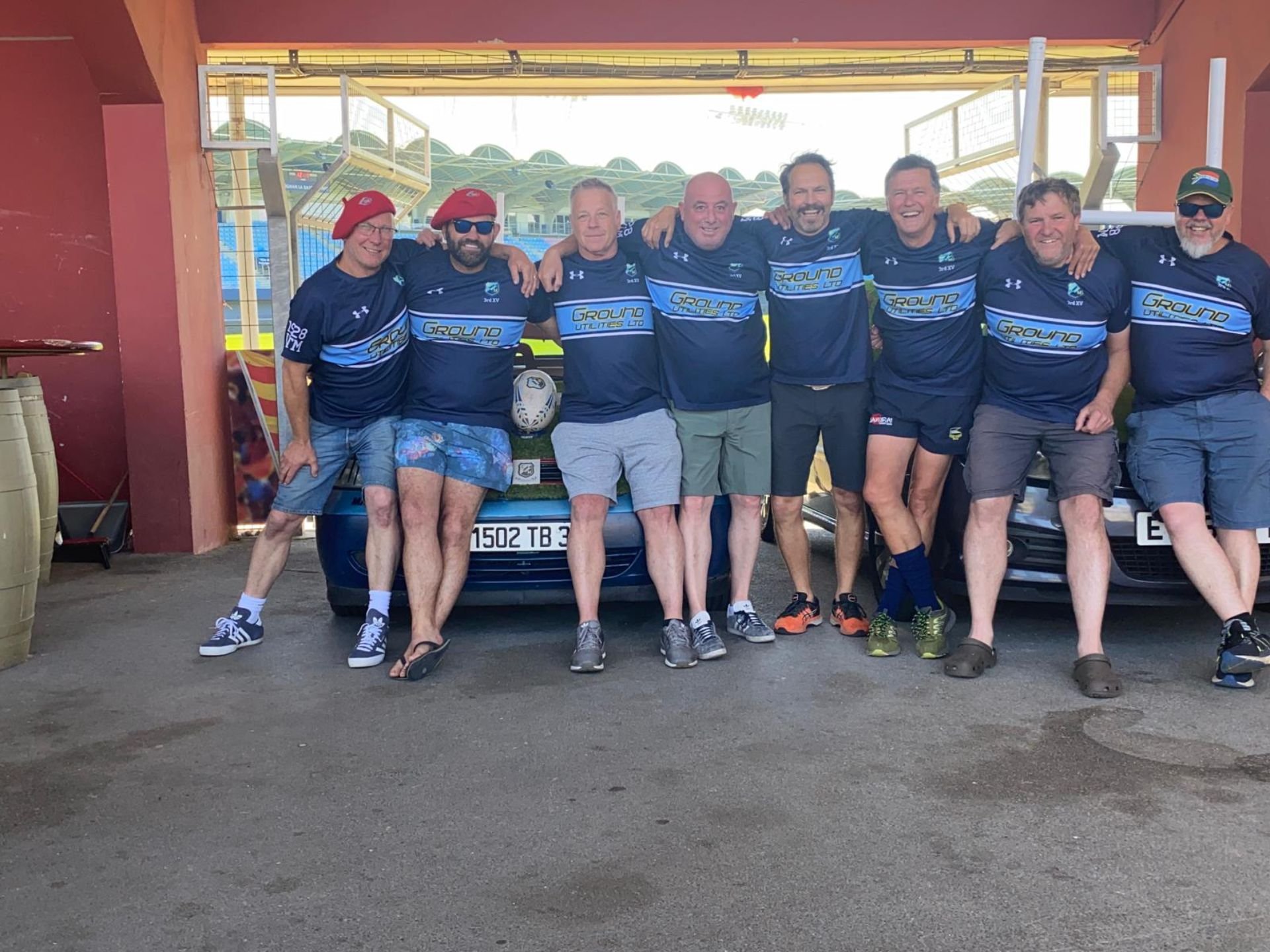 Group of eight men in sports jerseys standing together with arms around each other in front of two cars at a sports stadium or field, smiling and enjoying the moment.