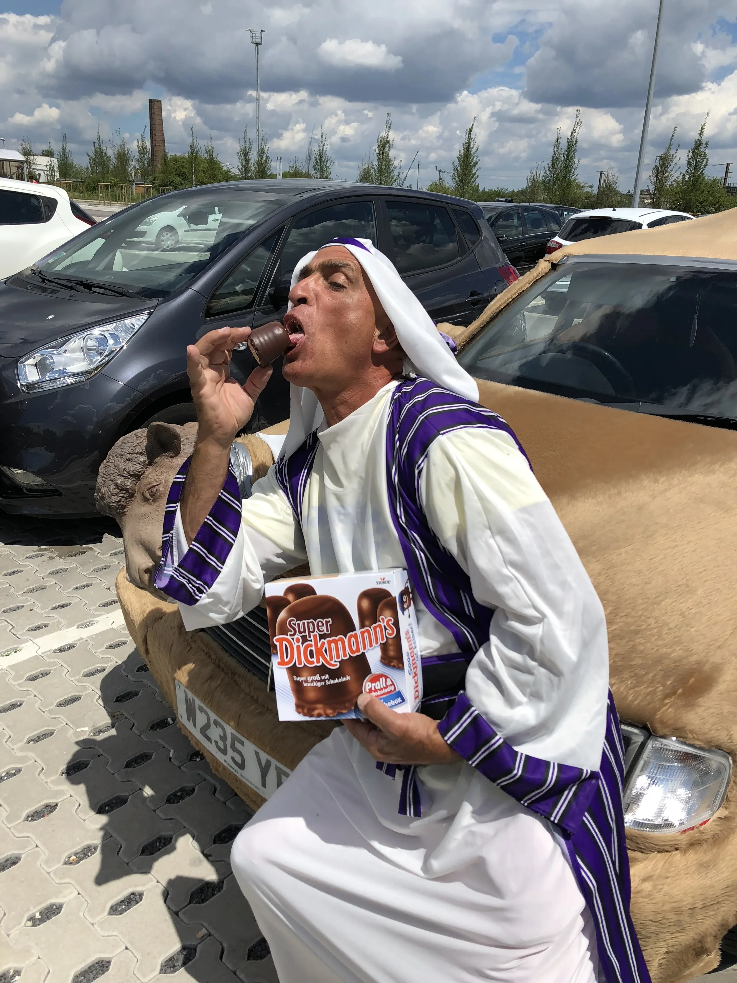 A man dressed in a costume resembling traditional Middle Eastern attire is sitting on the hood of a car, licking a chocolate candy. He is holding a box of Super Dickmann's chocolates. There is a camel behind him, and the scene appears to be in a park