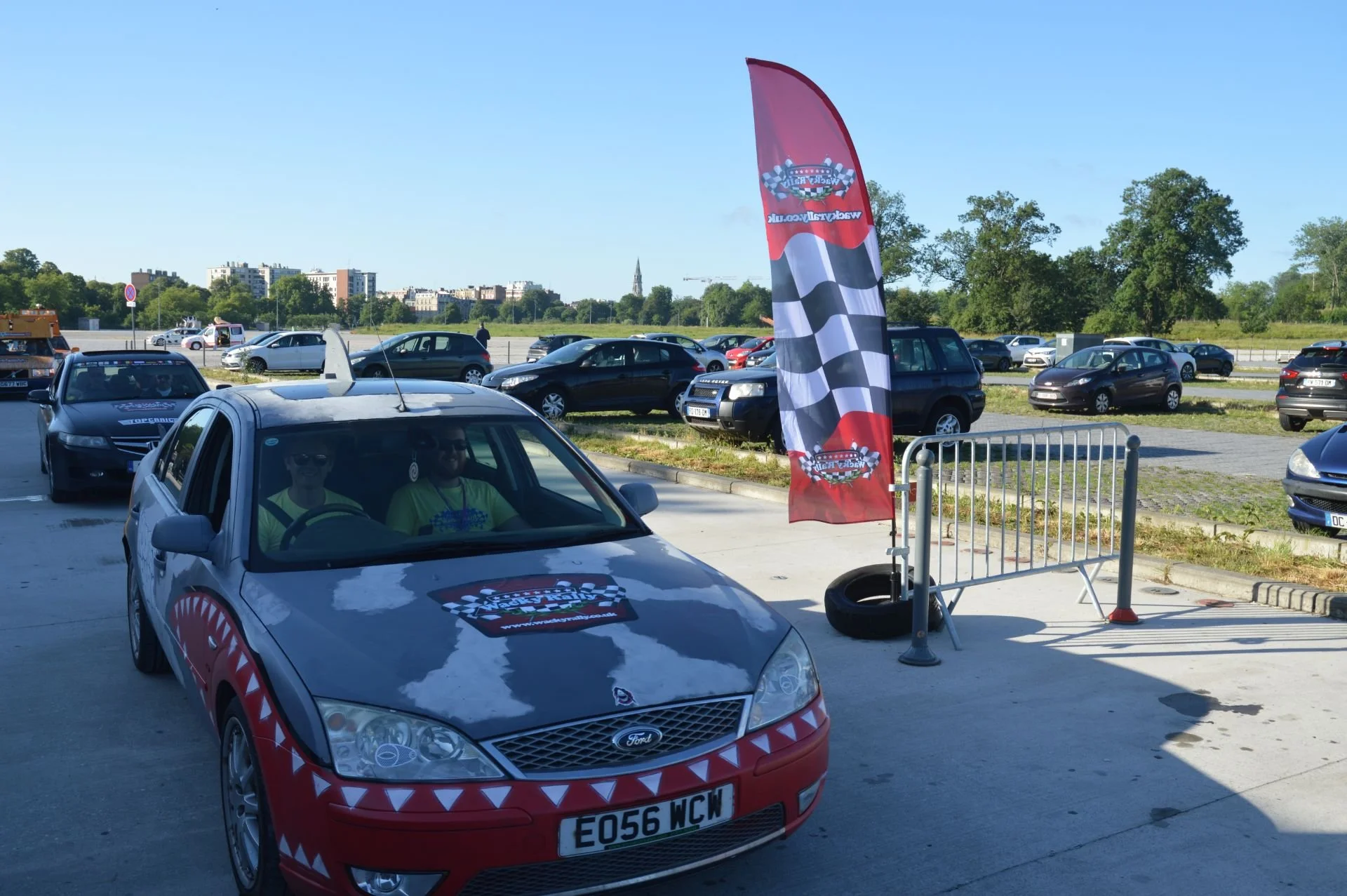A promotional event with a Ford car decorated with racing-themed graphics, parked in a lot with a checkered flag banner and several other cars in the background, under a clear blue sky.