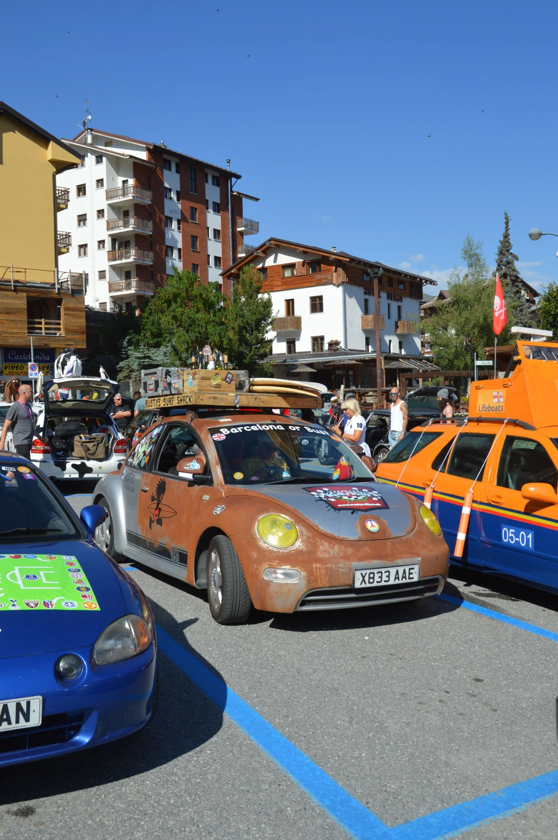 A parking lot filled with colorful cars, including a rust-colored Volkswagen Beetle, surrounded by people and buildings in a mountain town under a clear blue sky.