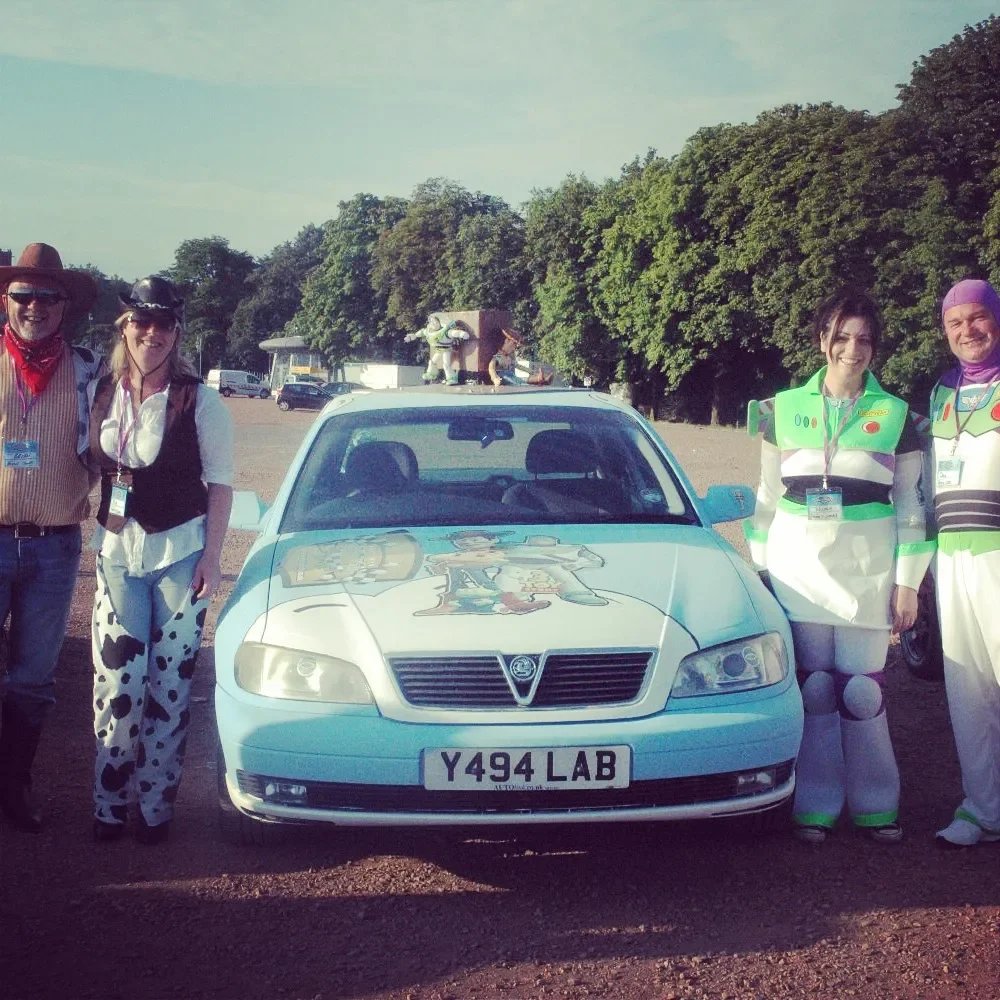 Group of five people dressed in costume costumes standing next to a decorated car with cartoon characters on the hood, in an outdoor parking lot with trees in the background.