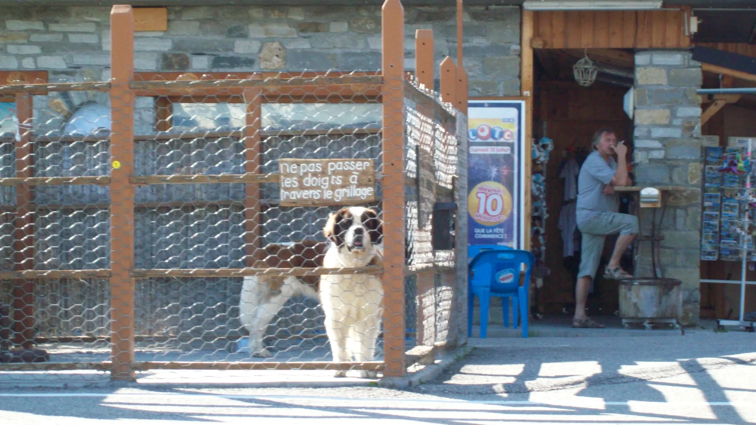 Dog inside a fenced enclosure with a sign that says not to pass fingers through the grill, a man sitting on a stool near a stone building with colorful posters and a man smoking a cigarette.