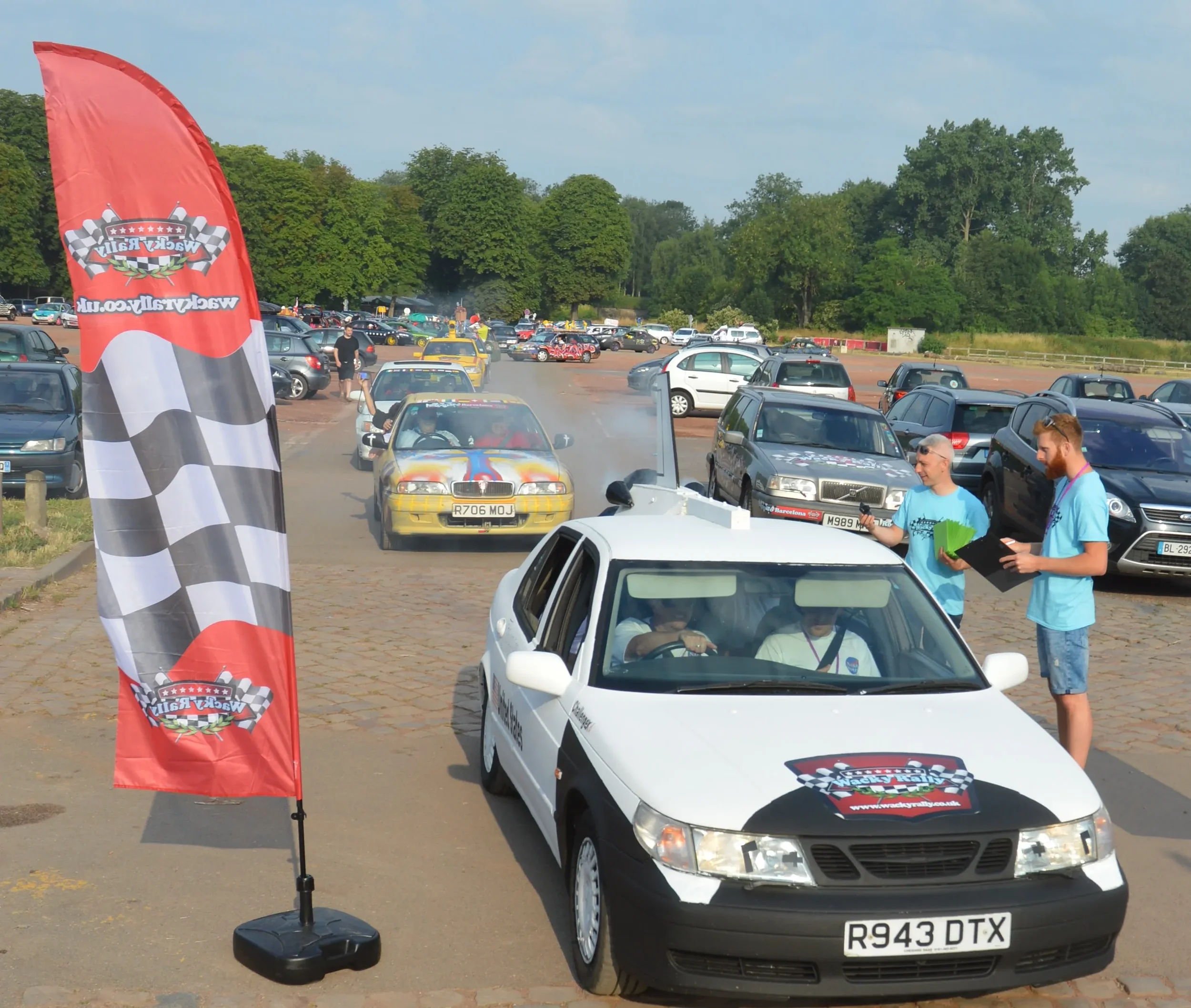 A white taxi with a rally logo on the hood, parked next to a flag with checkered pattern, at a car event. Two men in blue shirts are talking outside the taxi, one holding a clipboard and the other with a notepad. A yellow racing car with a rainbow de