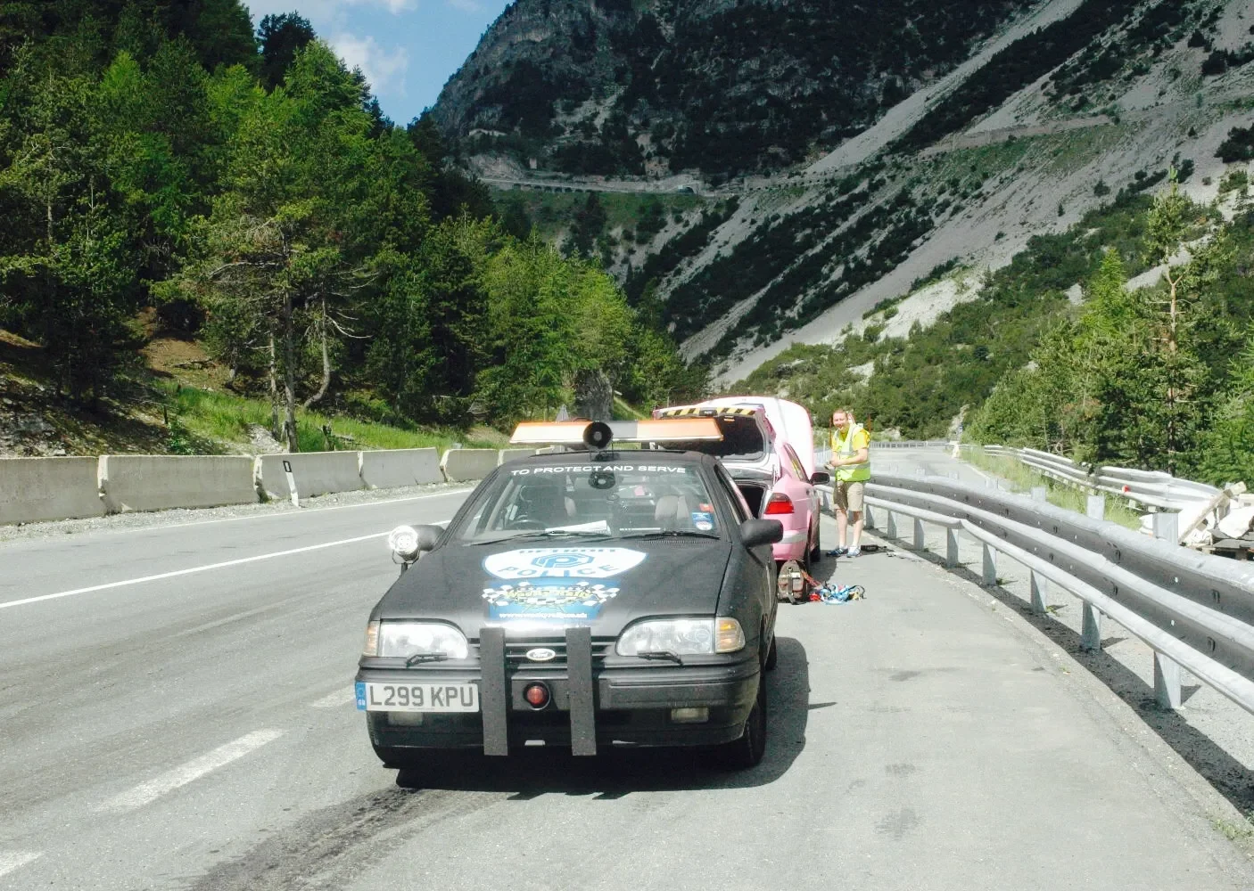 A police vehicle is stopped on the side of a mountain road with a man working behind the car near another stopped car. The background features forested mountains and clear sky.
