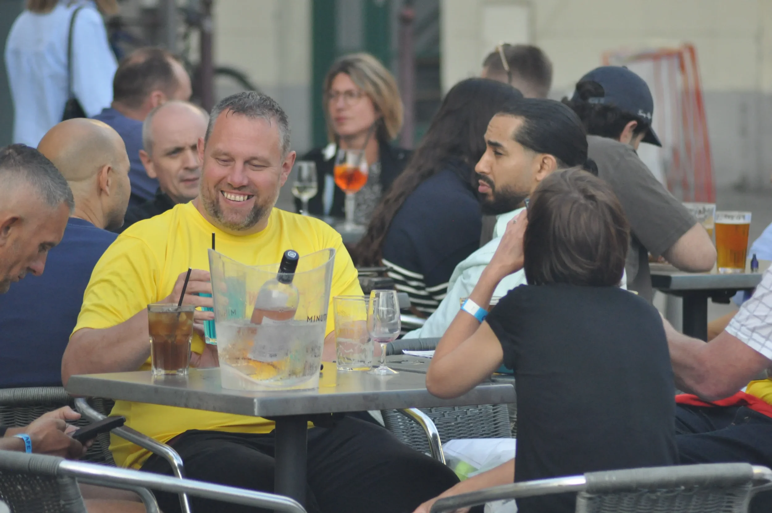 A group of people sitting at outdoor tables, drinking and socializing on a street patio, with some smiling and others engaged in conversation.