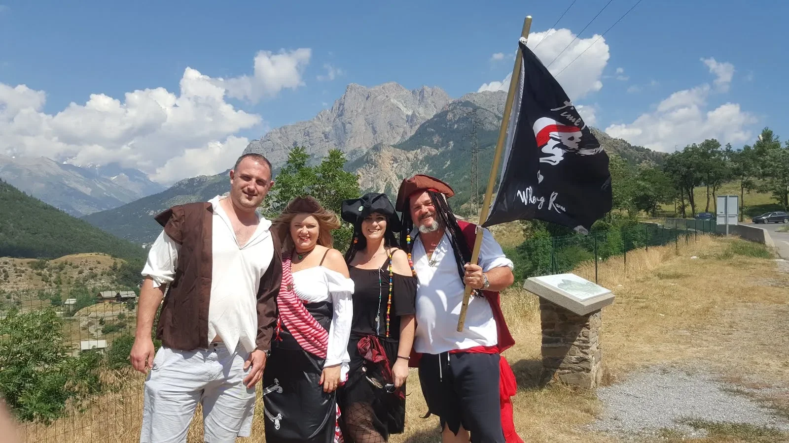 Four people dressed as pirates, standing outdoors with mountains in the background, holding a black pirate flag with a skull and crossbones.