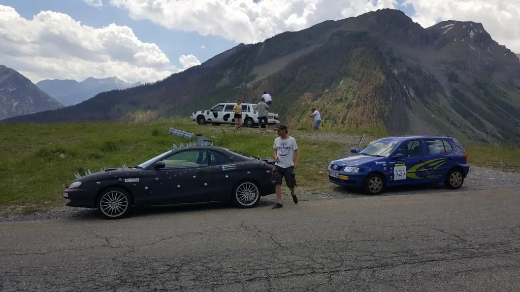 Two cars are parked on the side of a mountain road, with mountains and a cloudy sky in the background. Several people are near the cars, some standing and others sitting or looking at the scenery.