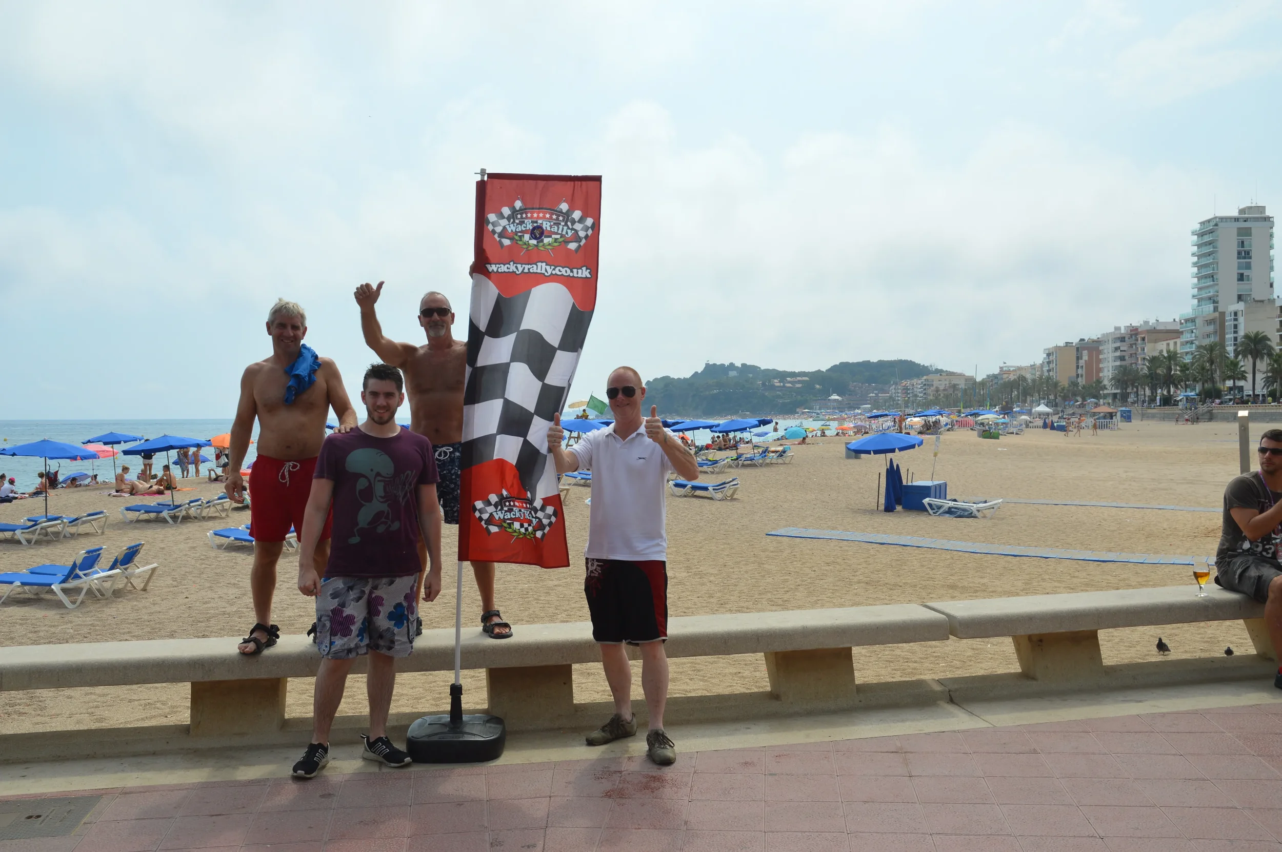 Four men on a beach posing with a racing flag, some giving thumbs up and smiling; colorful beach umbrellas and people in the background, along with buildings and trees.