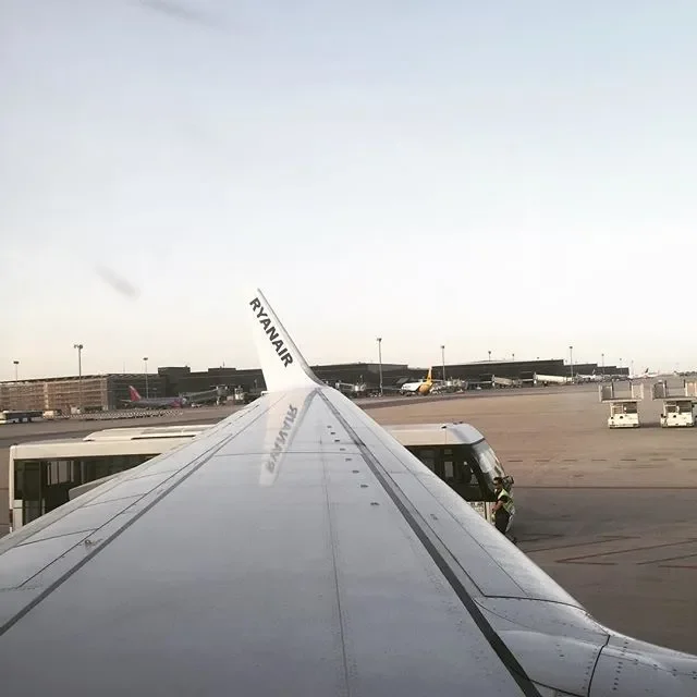 View from inside an airplane looking out over the wing at the airport tarmac with airport buildings and planes in the background during daytime.