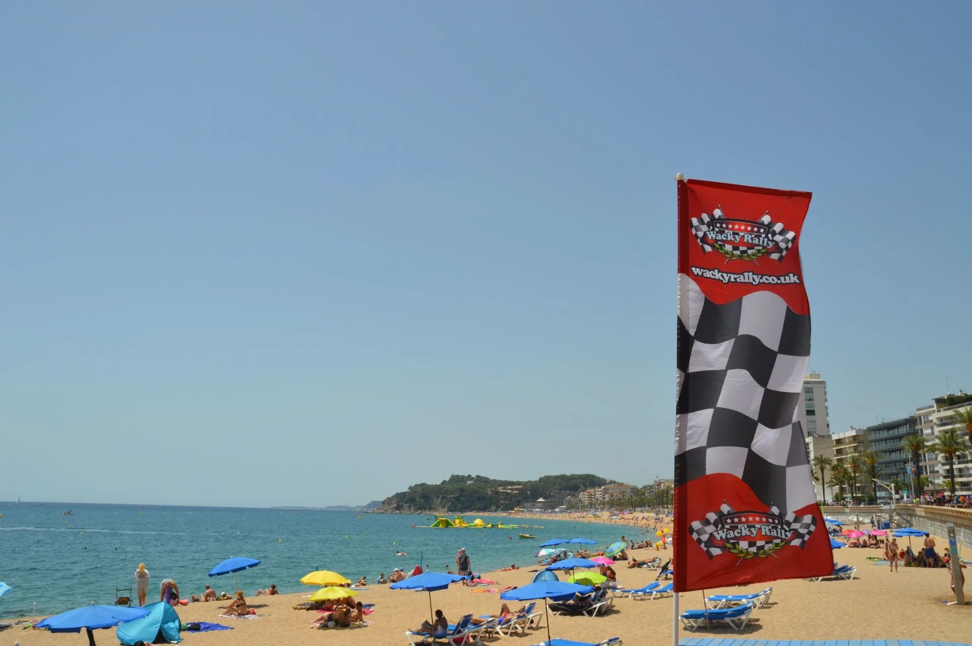 Beach scene with colorful umbrellas, people sunbathing, and a large flag with checkered patterns and the text 'Wacky Rally' on a flagpole, with buildings and palm trees in the background.