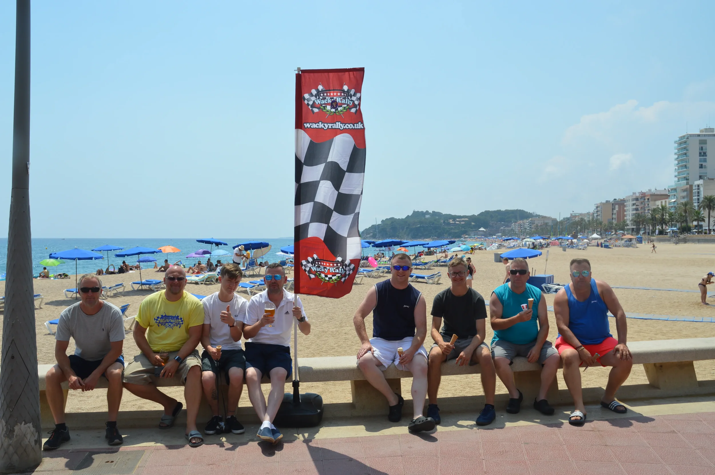 A group of nine men sitting on a beach bench with a wacky rally flag, beach umbrellas, and people in the background.