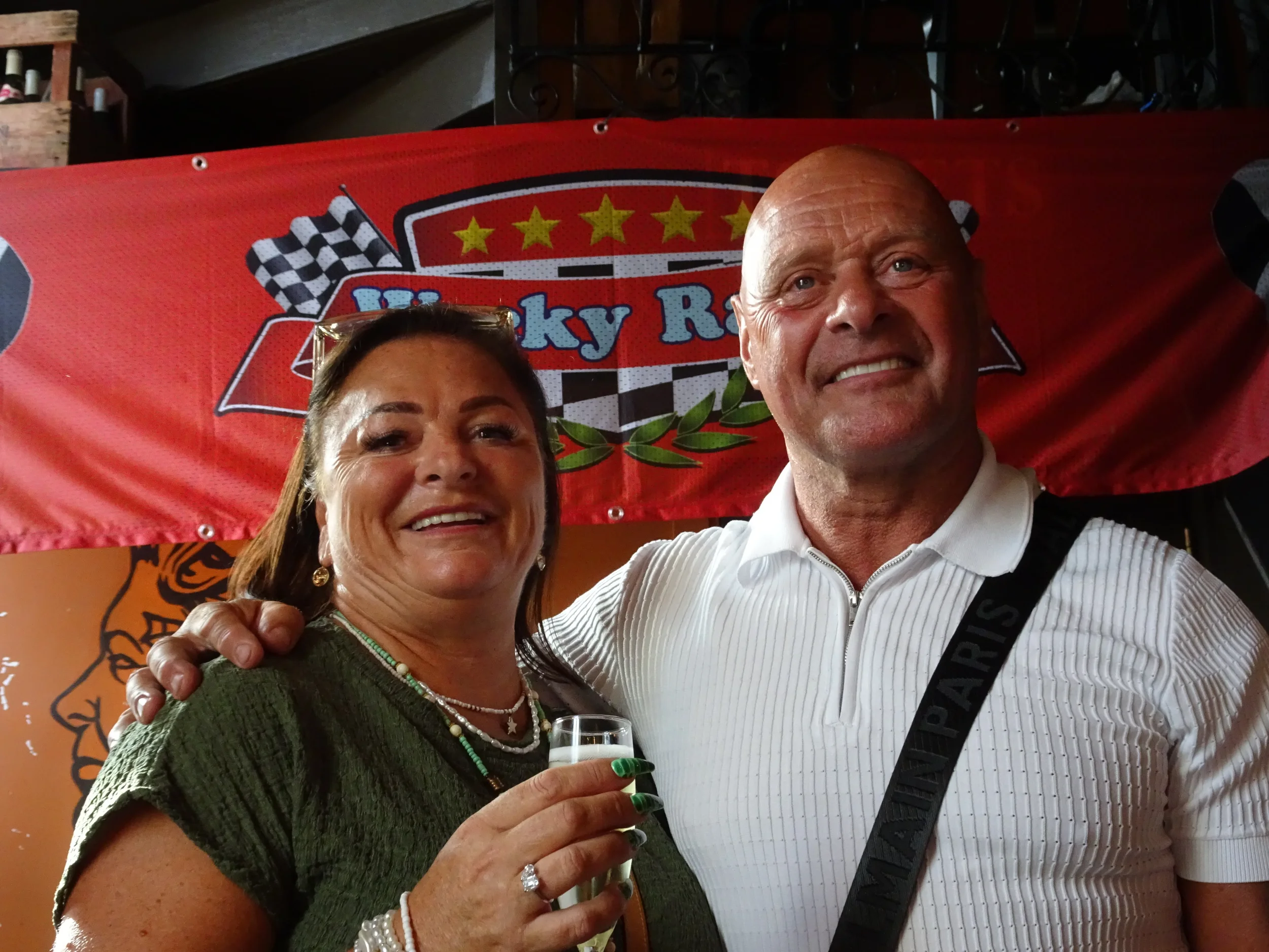 A smiling woman holding a champagne glass and a man with a black strap across his chest, standing in front of a red banner with racing theme, checkered flags, and the words 'Rocky R'.