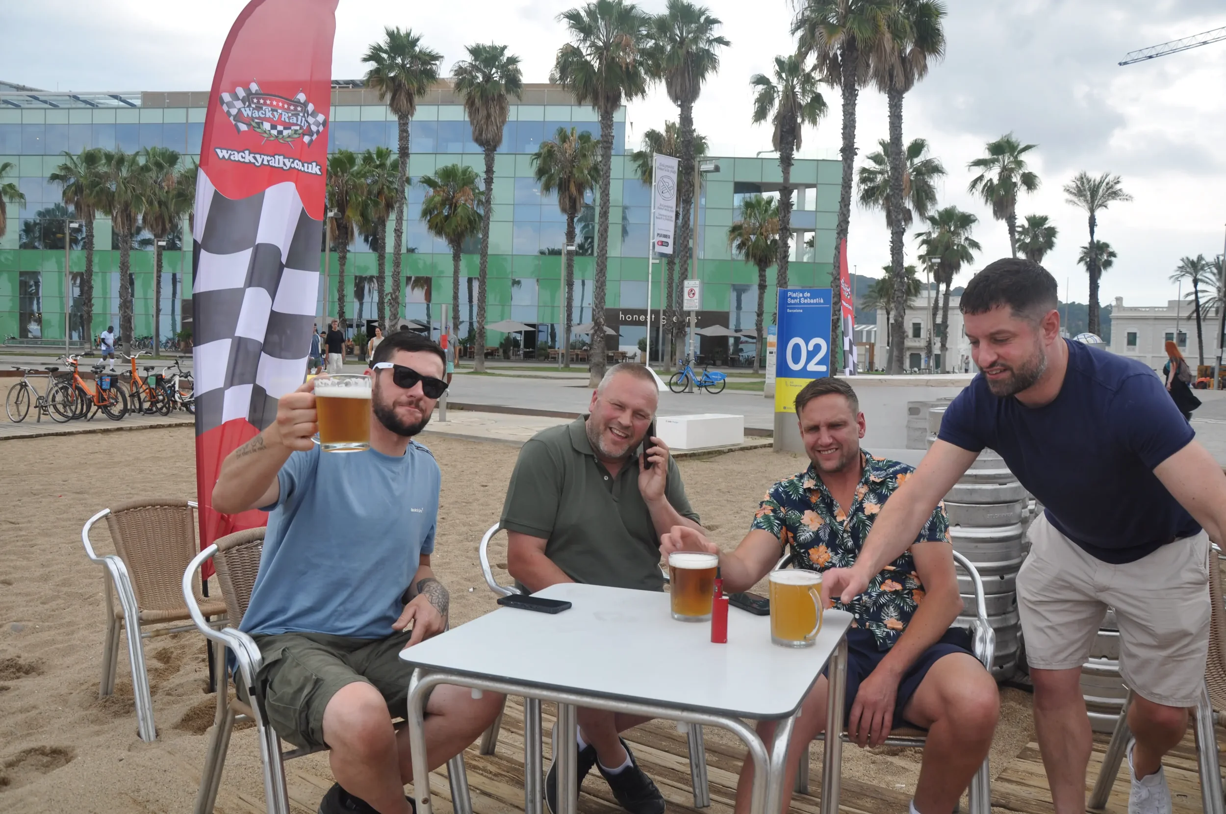Four men are sitting at a table on a beach, enjoying drinks and socializing. One man holds up a beer, another is talking on a cellphone, and the others are smiling. There are palm trees, bicycles, and a modern building in the background.