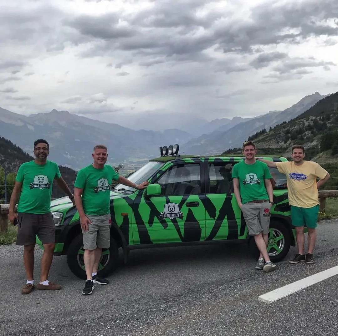 Five men posing next to a green and black striped SUV on a mountain road with cloudy skies and mountain scenery in the background.