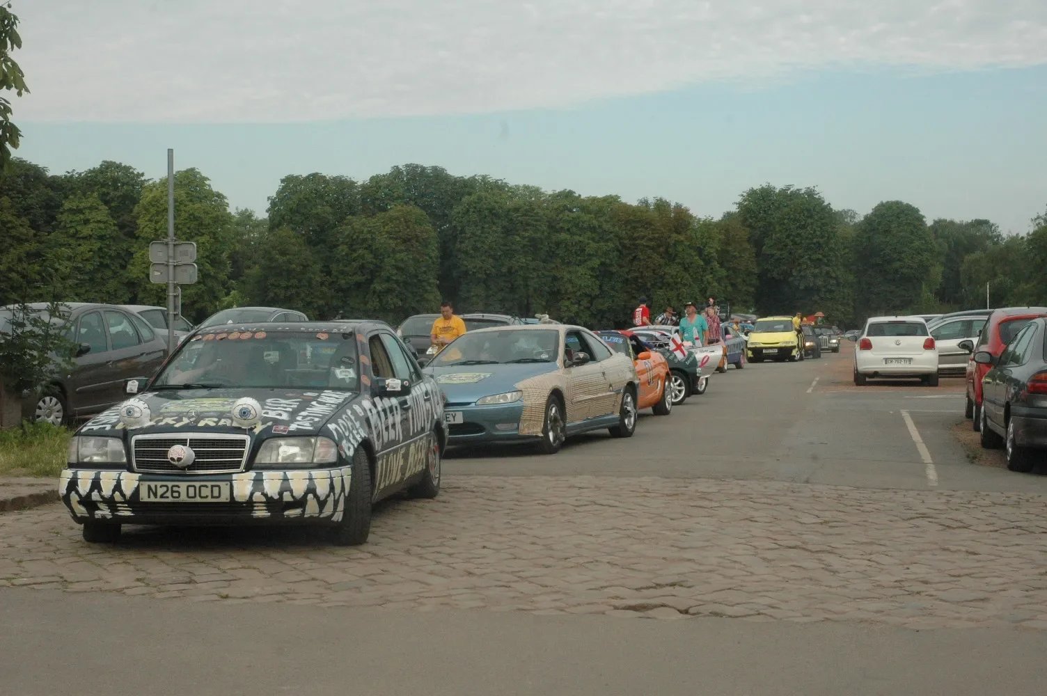 A parking lot with a line of decorated cars, including a black car with white graffiti-style writing and protective gear resembling eyes, and other cars with various themes and colors, with trees in the background.