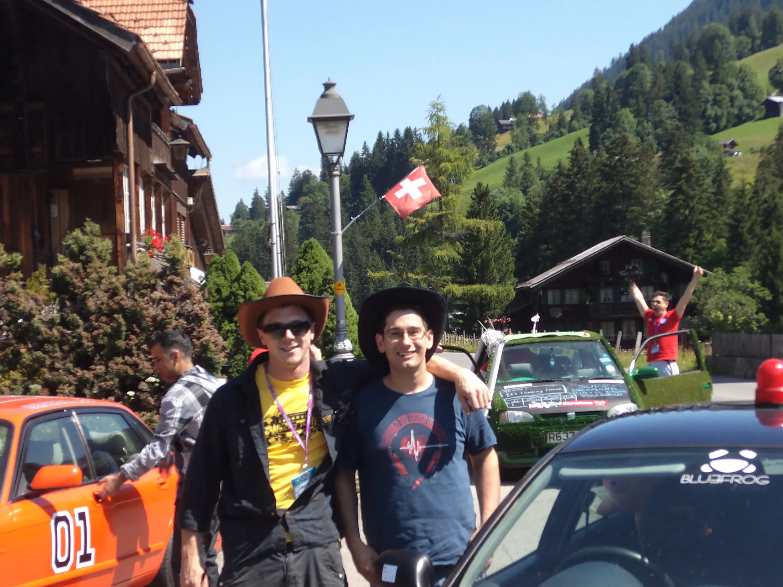 Two men smiling and posing with their arms around each other, standing in front of a row of cars in a mountainous area with houses and trees, while a young person in a red shirt stands next to a green car. A Swiss flag is flying in the background.