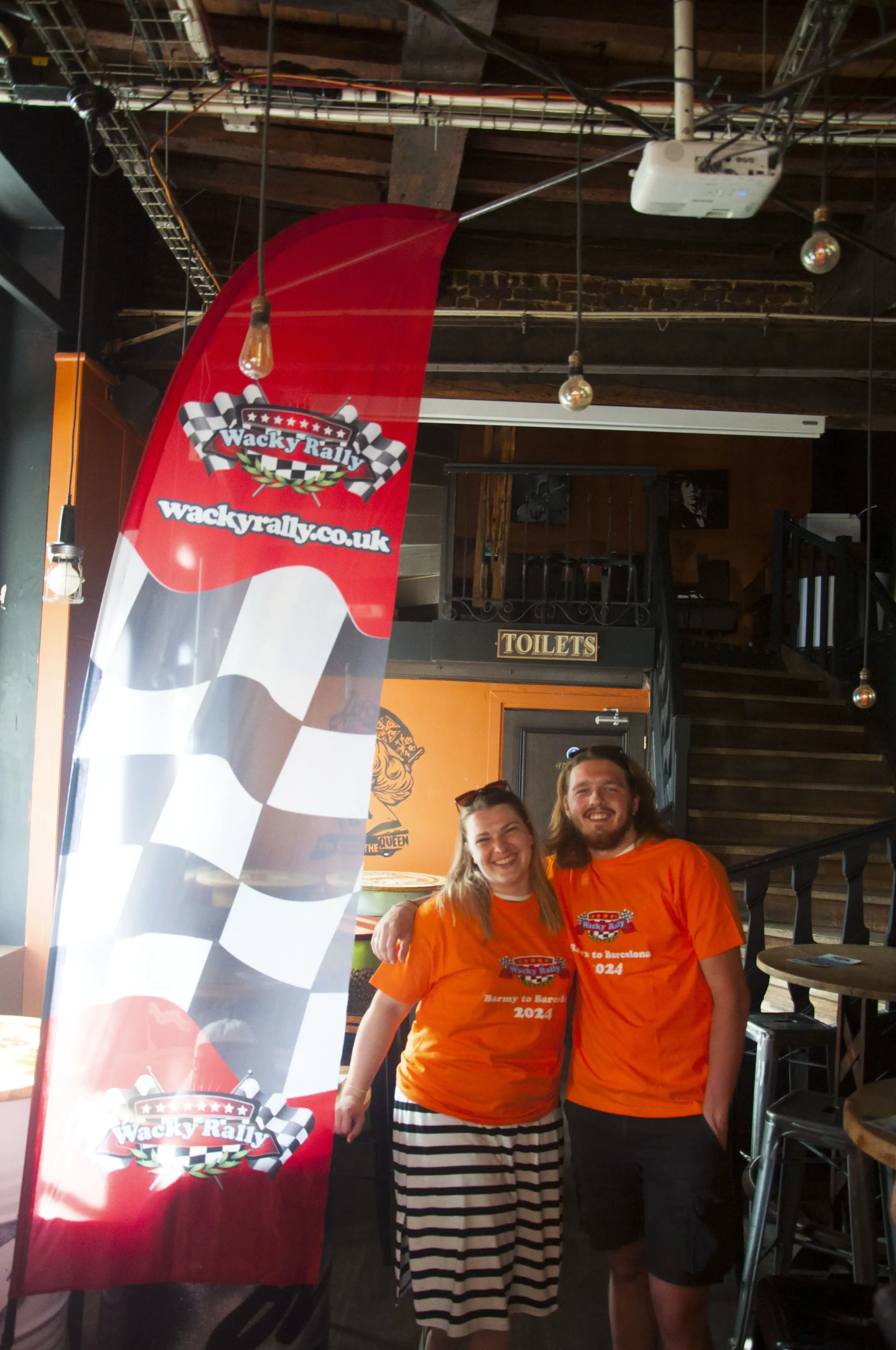 Two smiling people in orange T-shirts with racing flags logos, standing inside a venue with a Wacky Rally banner, brick walls, staircase, and a sign for toilets.